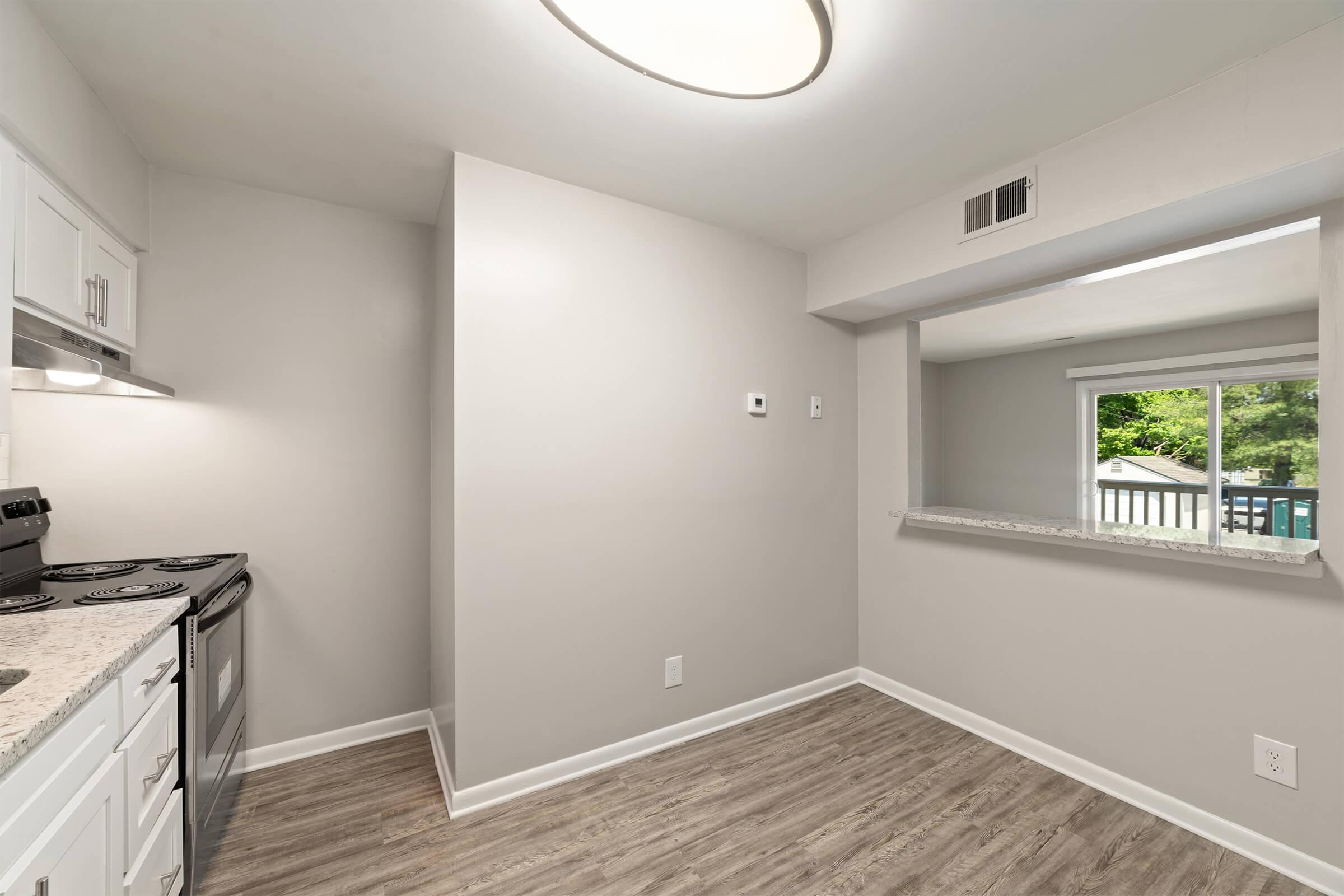 A modern kitchen with white cabinets and a granite countertop. The space includes a stove, and a window eases into an outdoor area, enhancing natural light. The walls are painted in a soft gray, and the flooring is a light wood laminate, creating a clean and contemporary look.