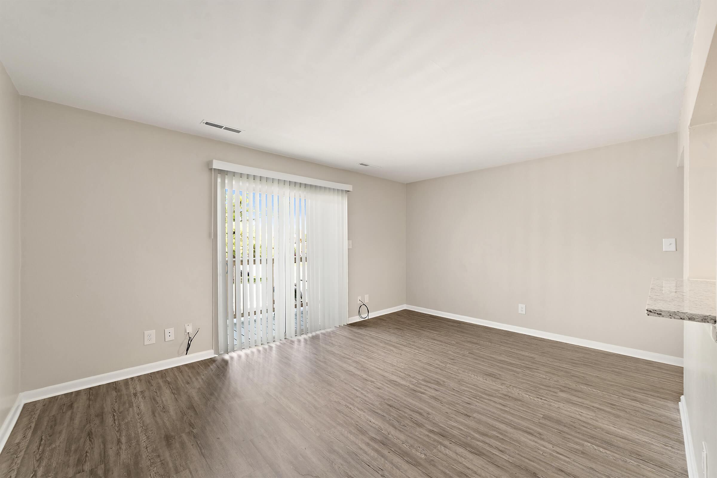 A spacious, empty room with light-colored walls and a large window covered by sheer white curtains. The floor is finished with light brown laminate, and the area appears bright and airy, suitable for various layout options.