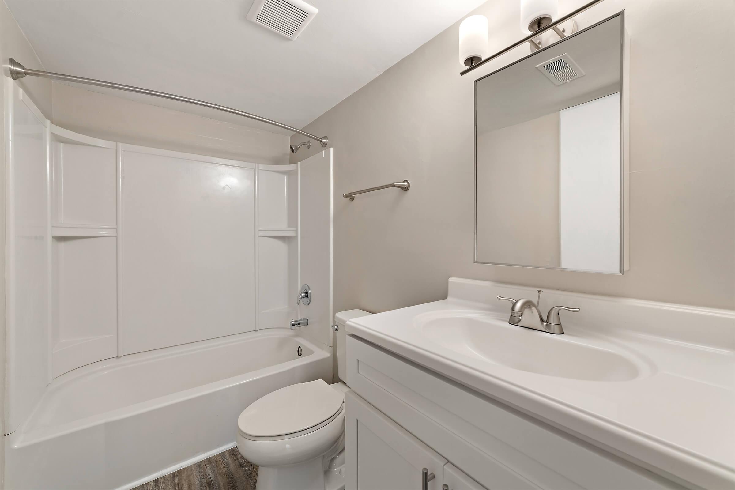 A clean and modern bathroom featuring a bathtub with a curved shower rod, a wall-mounted mirror above a white vanity with a sink, and a white toilet. The walls are painted a neutral color, and the flooring is a light wood. A ceiling vent is visible.
