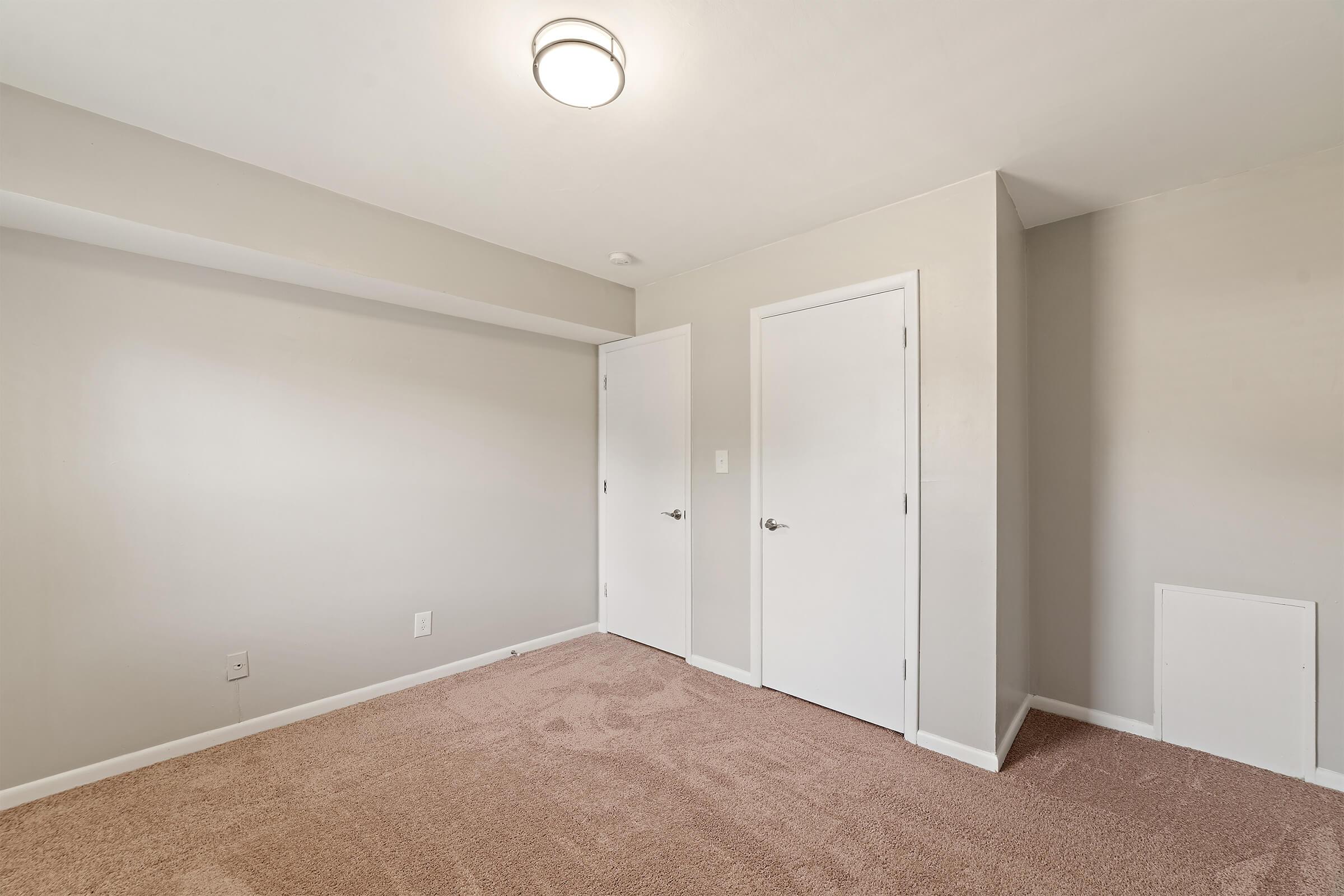 A spacious, empty room with light beige carpet. The walls are painted in a neutral tone, featuring two closed white doors on the right and a small wall vent. A ceiling light fixture illuminates the space, giving it a bright and airy feel. The room is devoid of furniture or decorations.