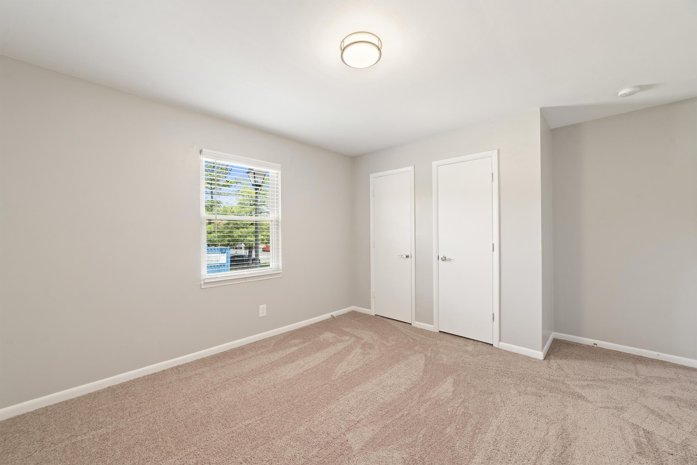 A bright, empty bedroom featuring light gray walls and plush beige carpet. It includes a window with blinds, providing natural light, and two white closet doors opposite the window. The ceiling has a modern light fixture, creating a clean and spacious ambiance.