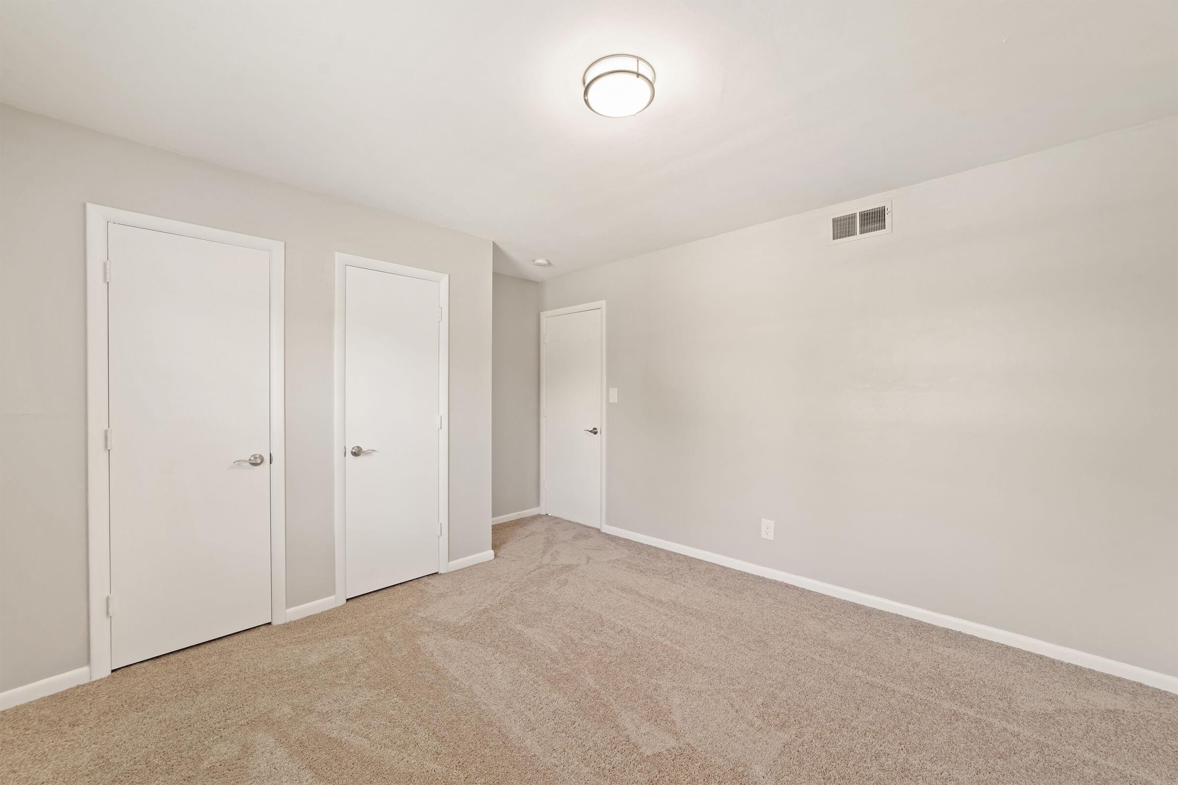 A spacious, empty room featuring light beige carpet, neutral walls, and a ceiling light fixture. Two closed white doors are visible on the left side, while a third door is on the right, leading to another space. The area is well-lit and designed for versatility in use.