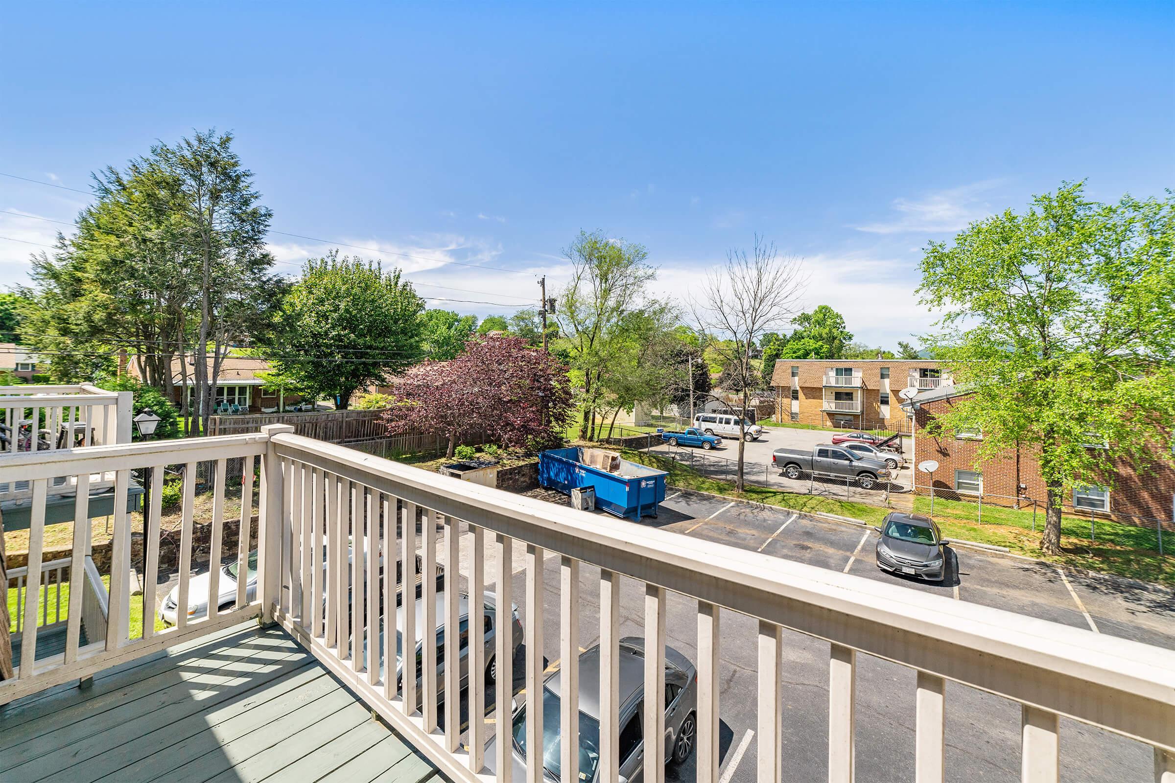 View from a balcony overlooking a parking lot and nearby trees, with residential buildings in the background. The sky is clear and blue, creating a bright atmosphere.
