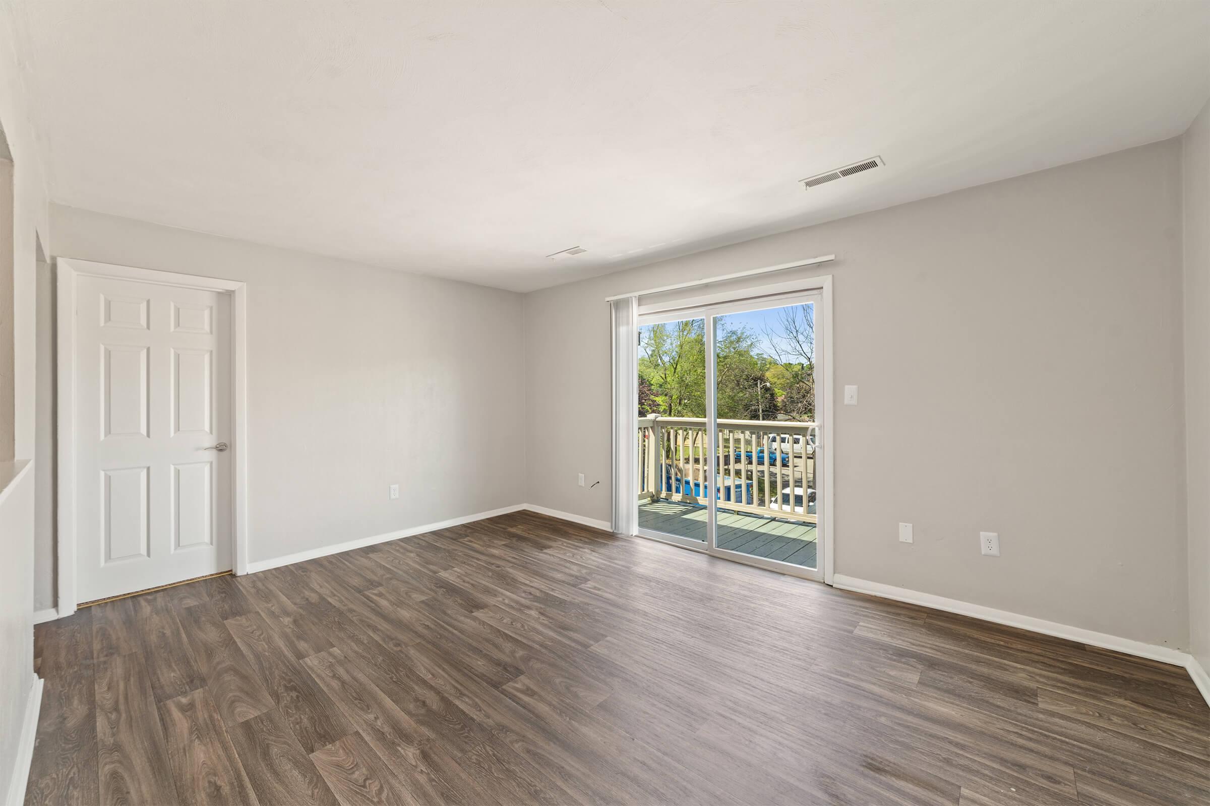 Empty room with light-colored walls and a wooden floor. There is a sliding glass door leading to a balcony, bringing in natural light and offering a view of greenery outside. A white door is visible on the left side.