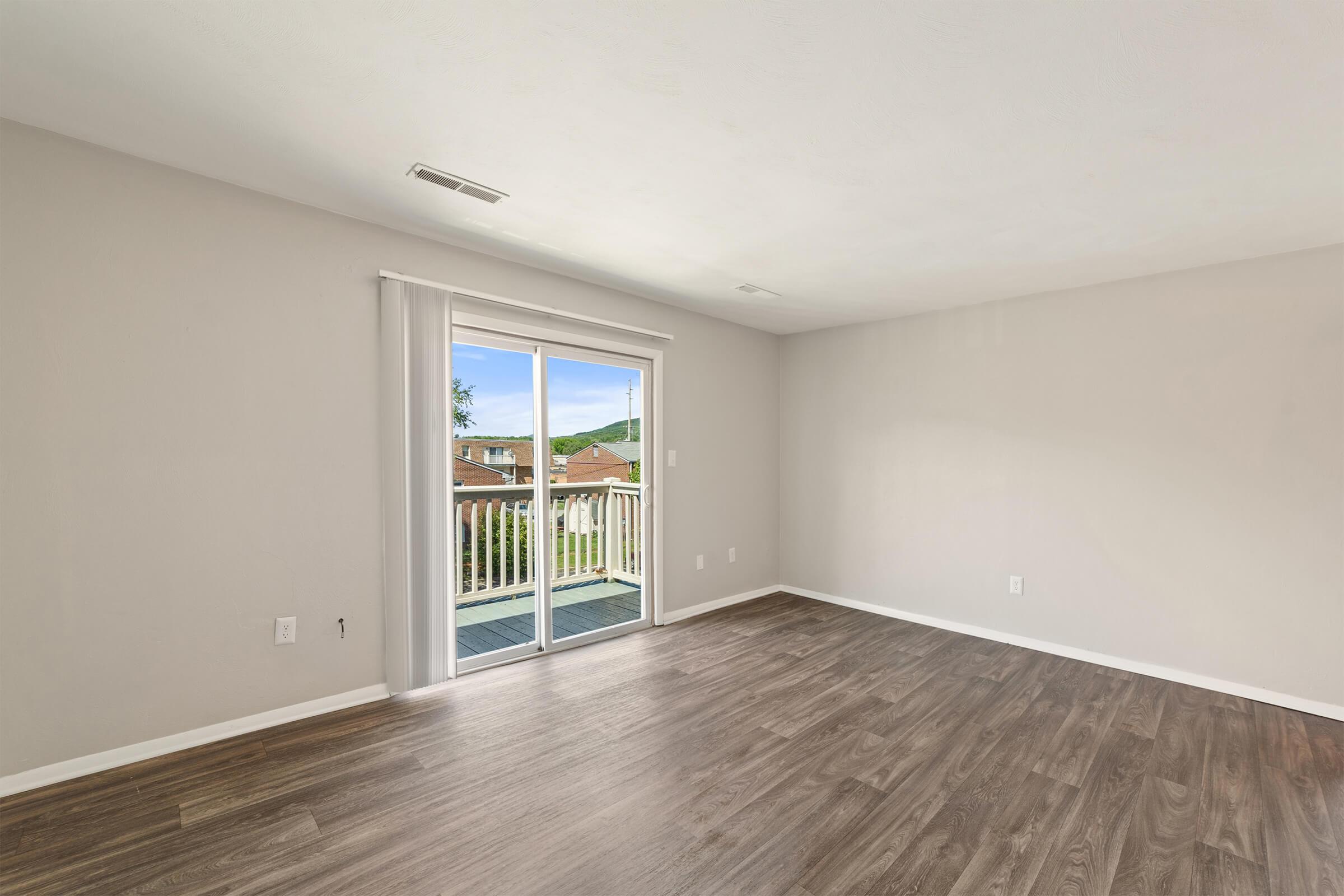A spacious indoor living area with a light-colored wall and wooden flooring. Large sliding glass doors provide access to an outdoor balcony, allowing natural light to fill the room. The view outside shows a pleasant landscape with greenery and rooftops in the distance.