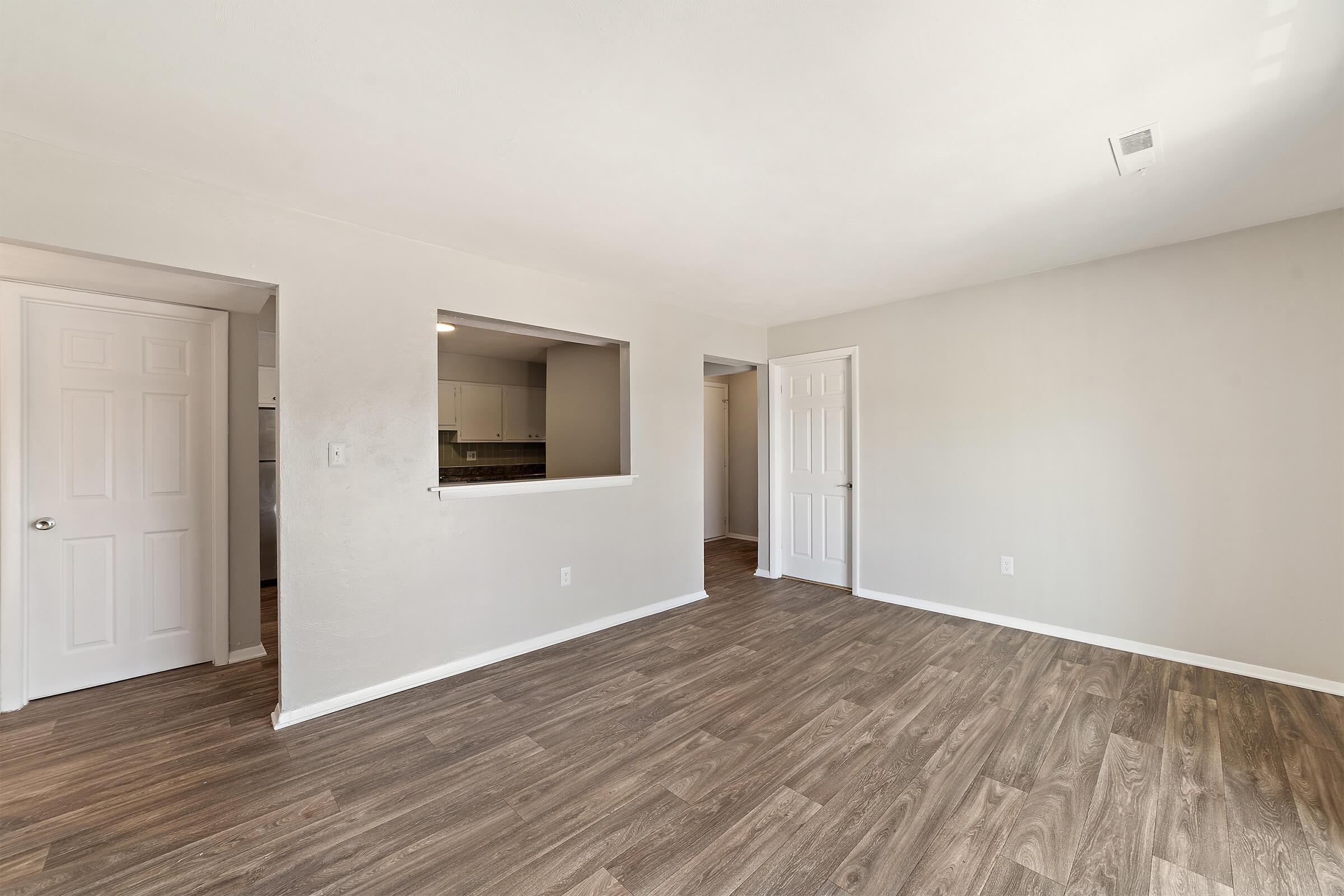 A well-lit, empty living room with light-colored walls and wooden flooring. There are two doorways, one leading to a kitchen area and the other to a hallway. The space has a modern feel, with a slight open concept and no furniture or decor. Natural light enters through the windows, enhancing the spaciousness.