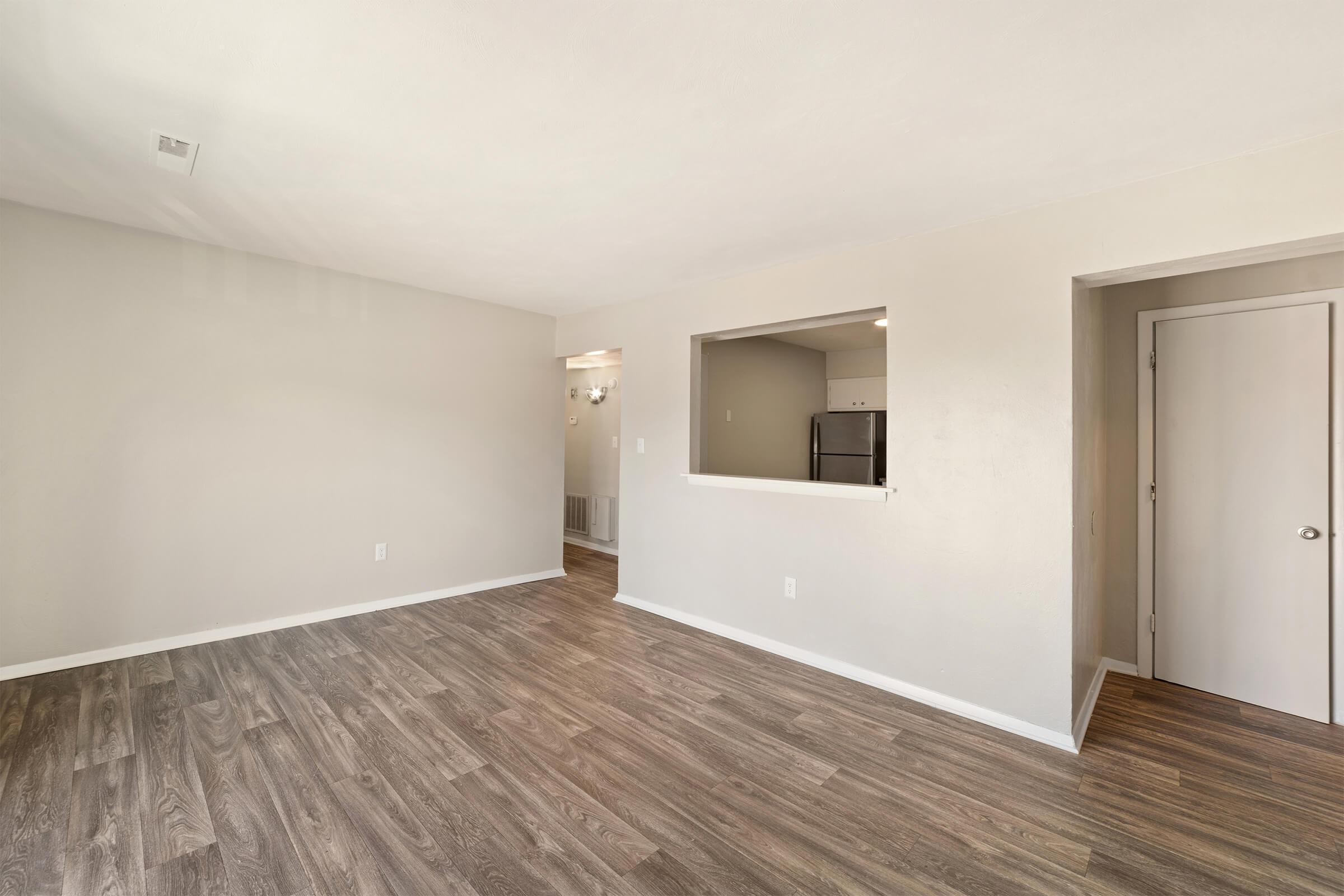 A spacious, empty room with light-colored walls and wood-like flooring. There is an opening leading to a kitchen area in the background and a doorway on the right. Natural light illuminates the space, creating a bright and airy atmosphere.