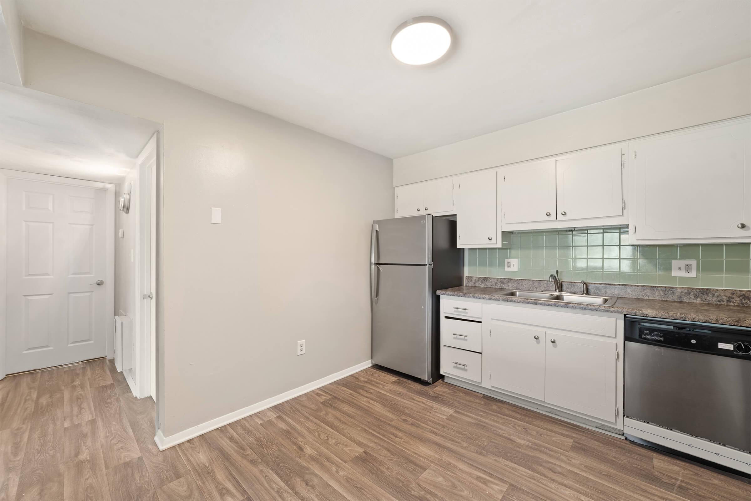 A modern kitchen featuring light-colored cabinets, stainless steel appliances including a refrigerator and dishwasher, a sink with countertop space, and green tiled backsplash. The kitchen opens up to a hallway with a door leading outside, and the floor has a wood-like appearance. Bright overhead lighting enhances the space.