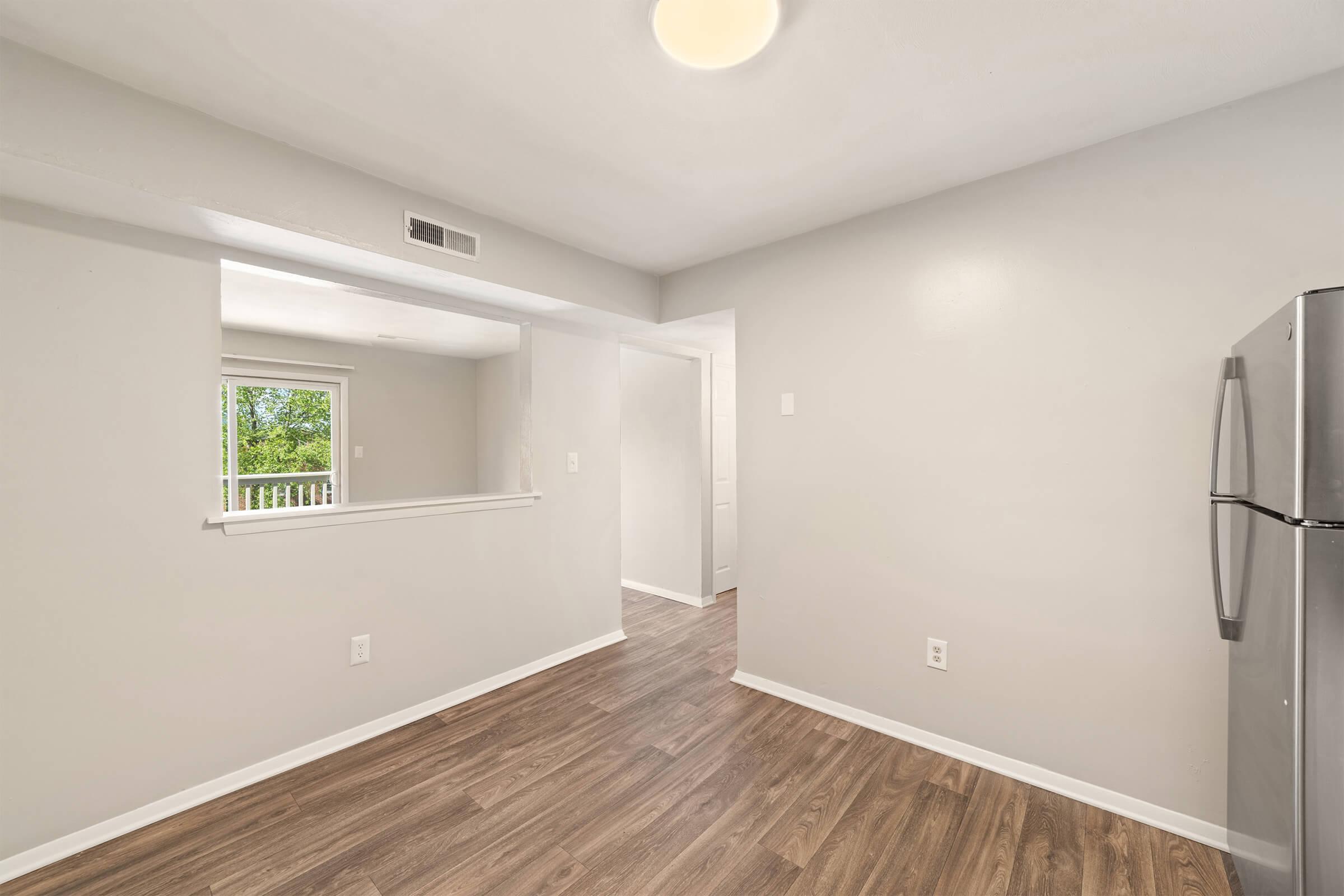 A modern kitchen space with light gray walls and hardwood flooring. It features a view of an adjoining room and a window allowing natural light, along with a stainless steel refrigerator visible on the right. The overall atmosphere is bright and inviting, suitable for culinary activities.