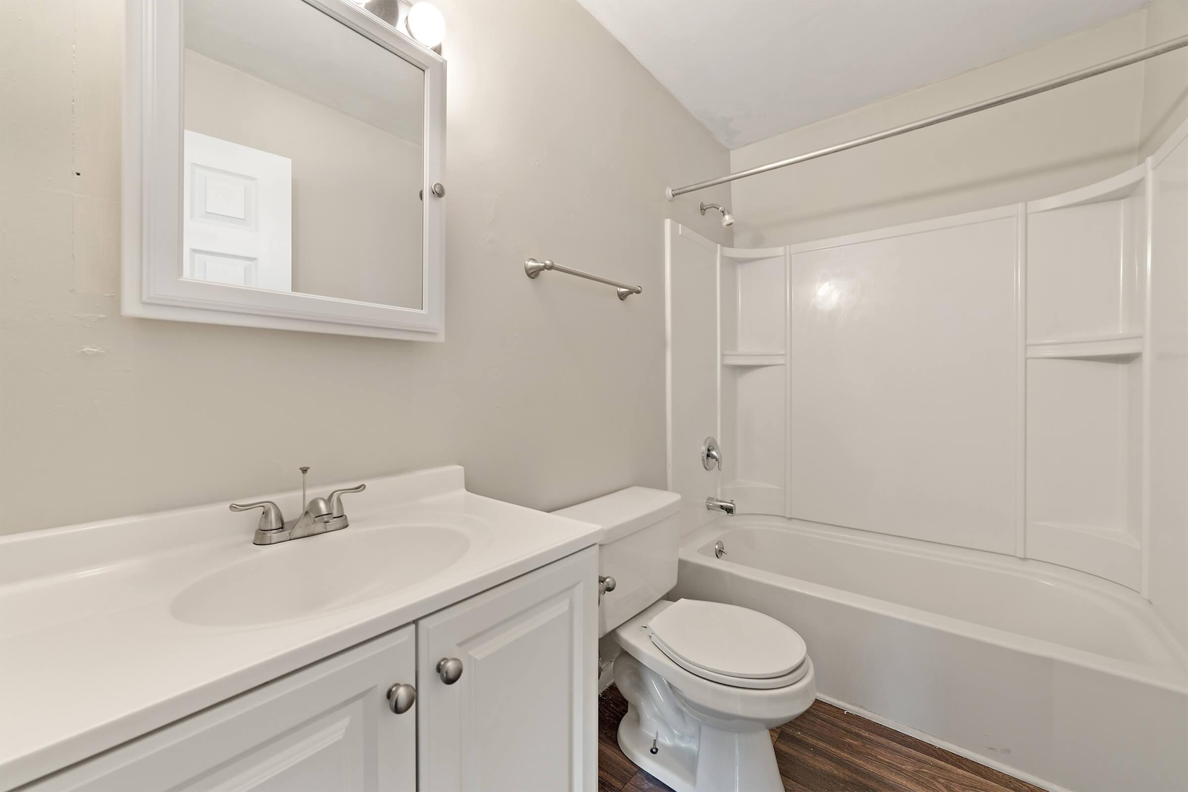 A clean, modern bathroom featuring a white sink with cabinet storage, a large mirror, a toilet, and a bathtub with a shower. The walls are painted in a light color, and the flooring is a warm wood finish. The overall design is simple and functional.