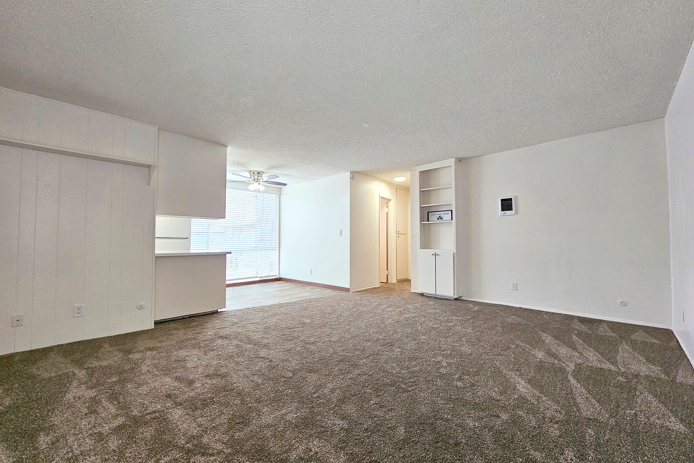 A spacious living area featuring plush, light brown carpet, white walls, and a ceiling fan. To the left, there’s an open kitchen with a bar area. Natural light enters through a window, creating a bright atmosphere. The room has a cozy, inviting feel with minimal furniture.
