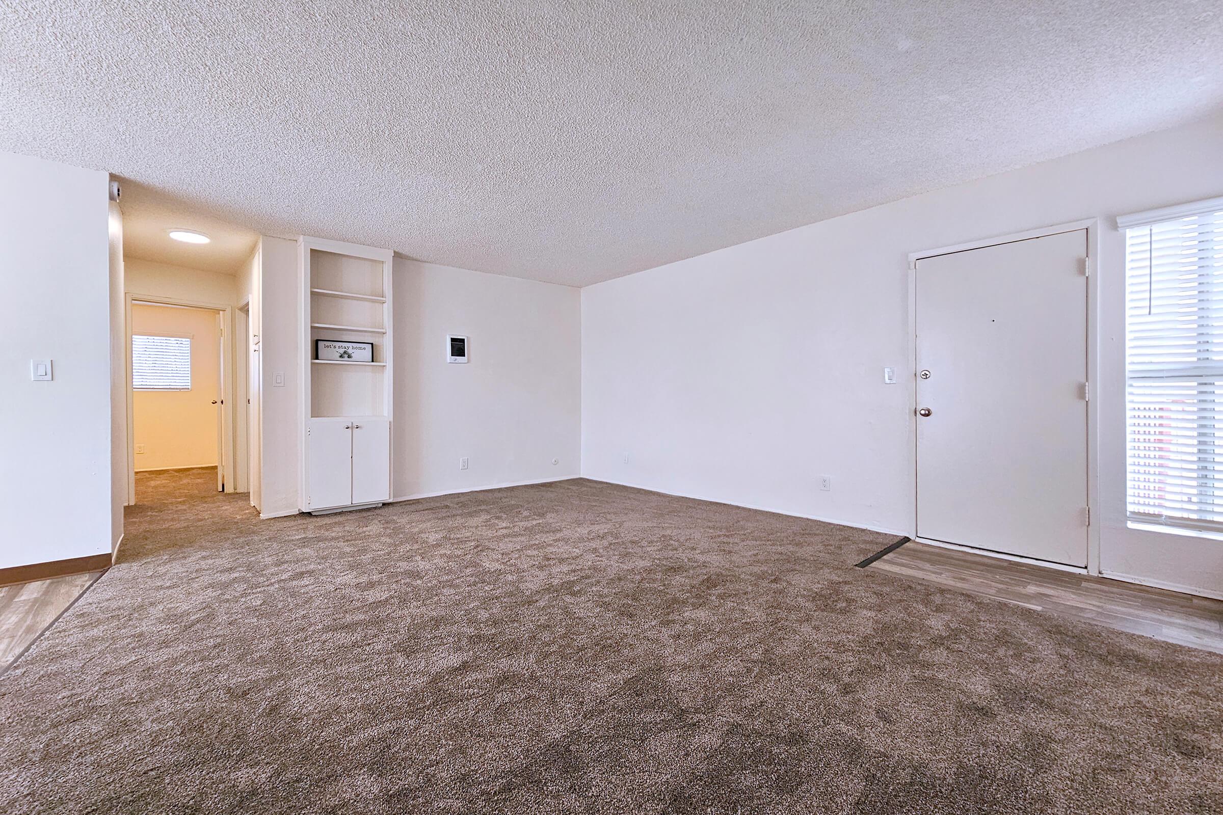 A bright and spacious room featuring light-colored walls and carpet, with a doorway leading to another room. On the left, there is a built-in shelf or cabinet, and a front door on the right. Natural light streams in from a window, creating an inviting atmosphere.