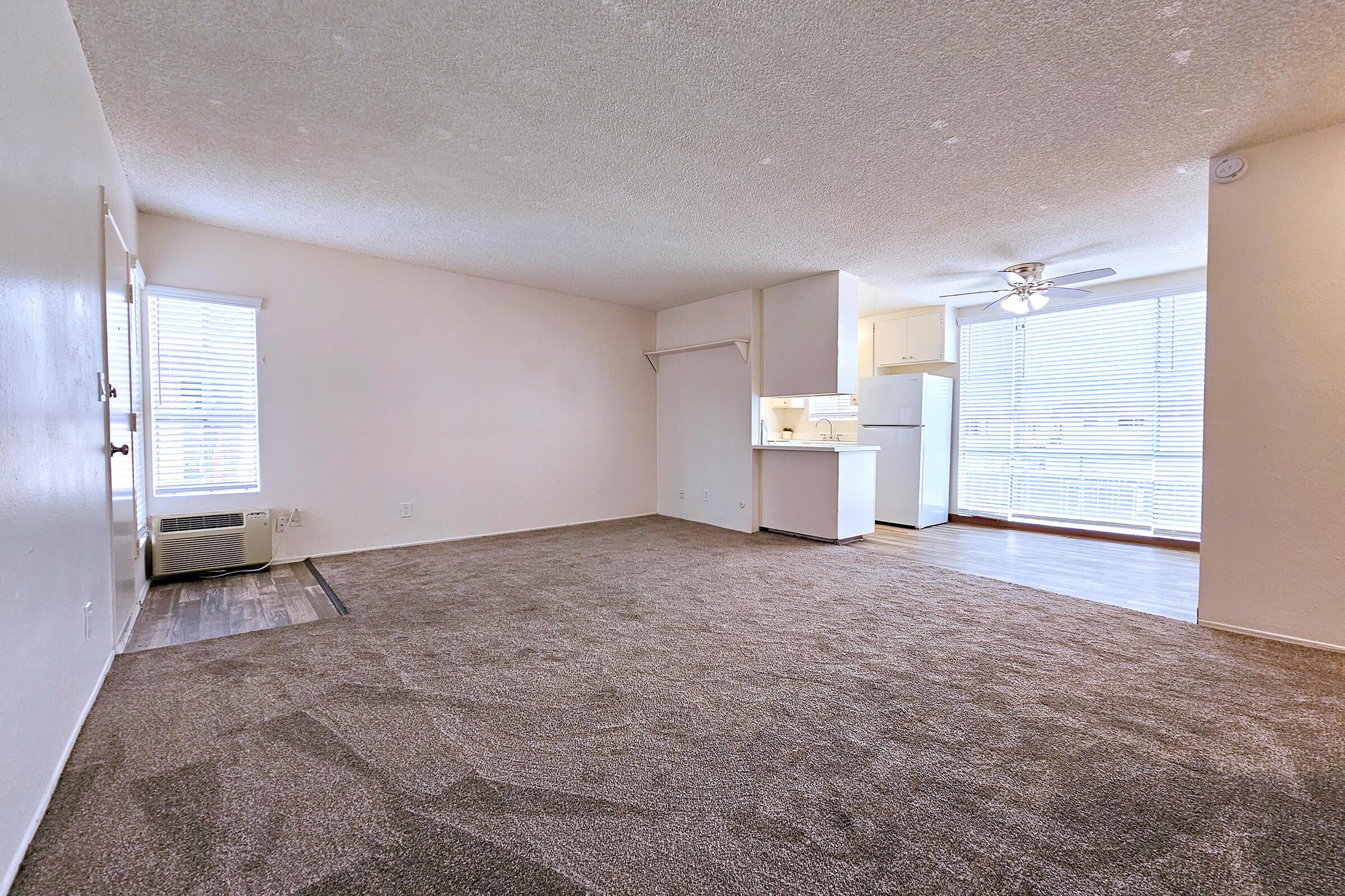An empty living room showcasing a spacious layout with light-colored walls, carpeted flooring, and a ceiling fan. There is a small kitchenette visible in the background, illuminated by natural light from large windows. The room is designed to create a warm and inviting living space.