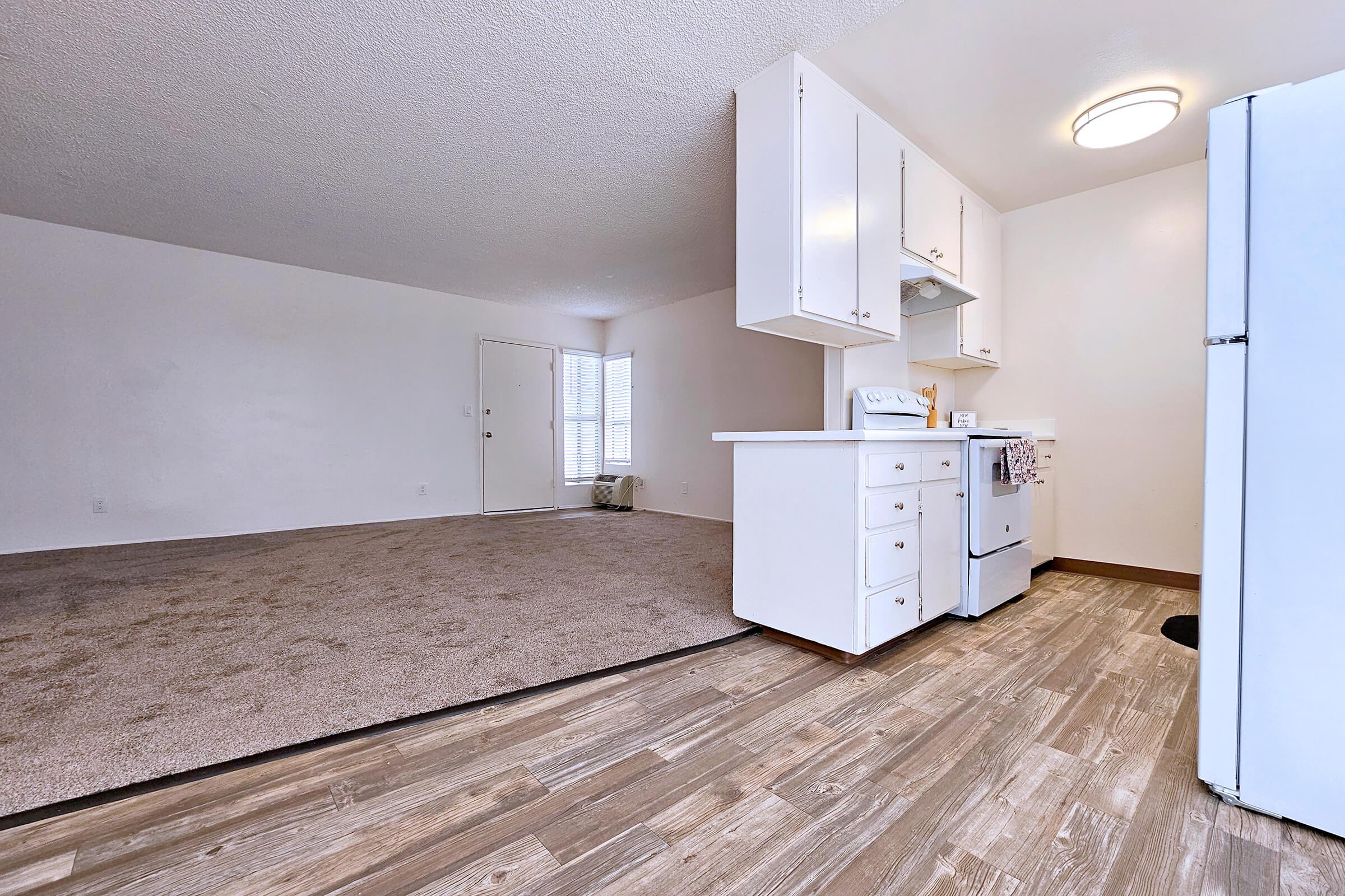 A spacious, well-lit apartment interior featuring a living area with beige carpet and a kitchen area with white cabinets and appliances. A door leads outside, and there are large windows allowing natural light in. The flooring is a modern wooden design, adding warmth to the space.