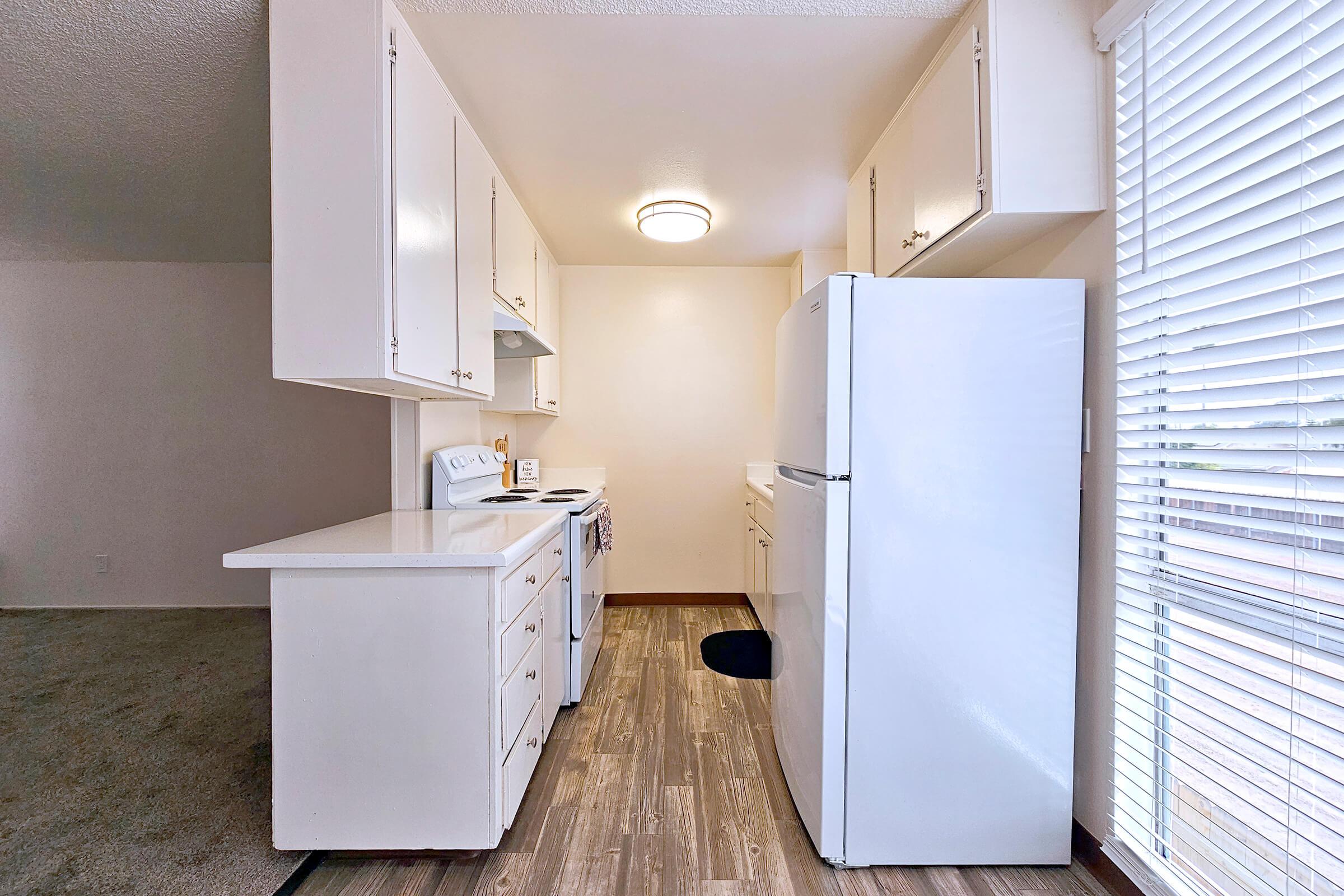 A bright kitchen featuring white cabinets, a white refrigerator, and a stove with an oven. The countertop is clear, and there's a window with blinds letting in natural light. The flooring is wood-like, and there is a small area rug on the floor.