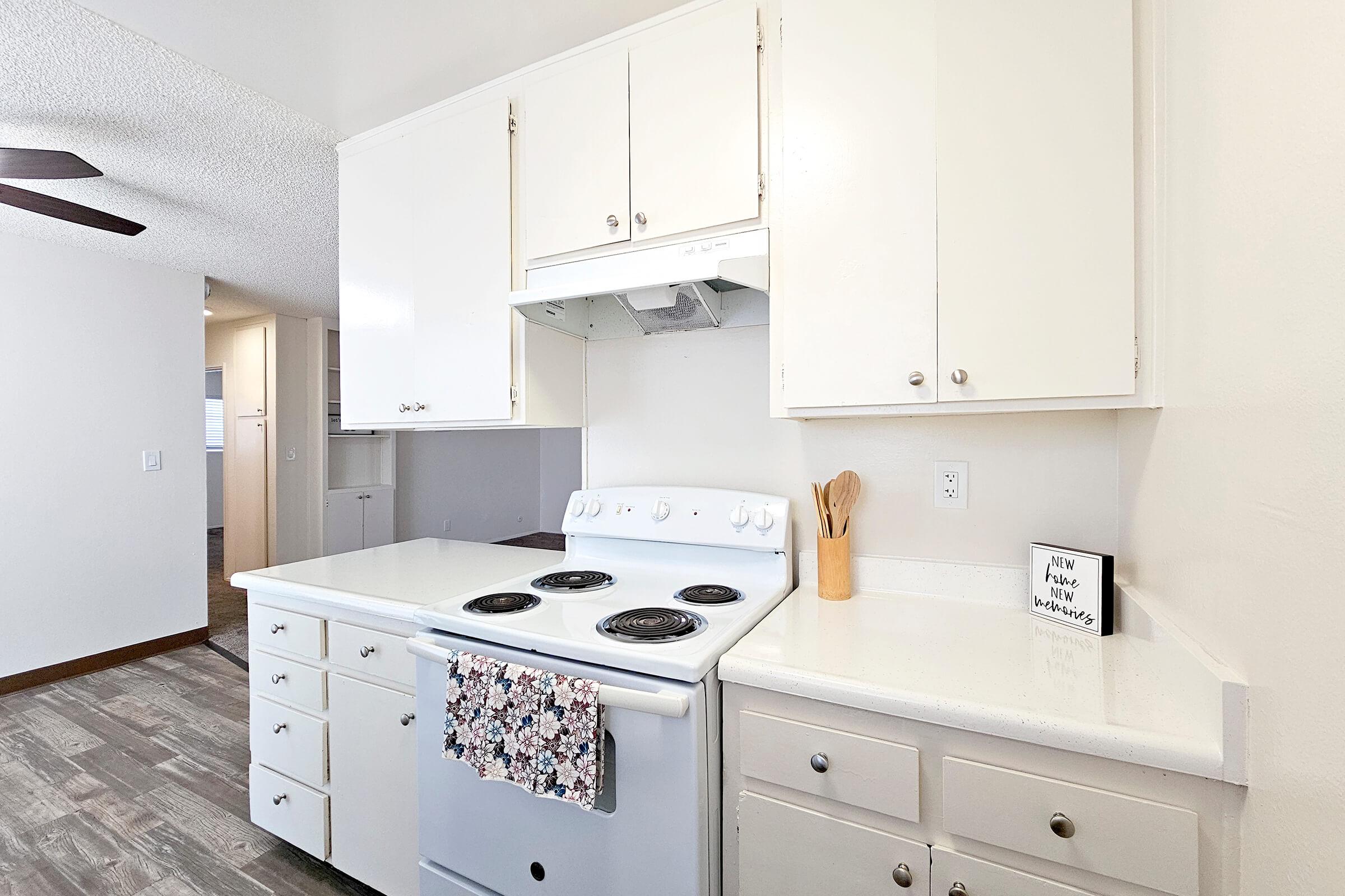 A clean and bright kitchen featuring white cabinetry, a white countertop, and a stovetop with four burners. There is a wooden utensil holder on the countertop and a decorative dish towel hanging from the stove. The background shows an open space, giving a glimpse of a connected living area.