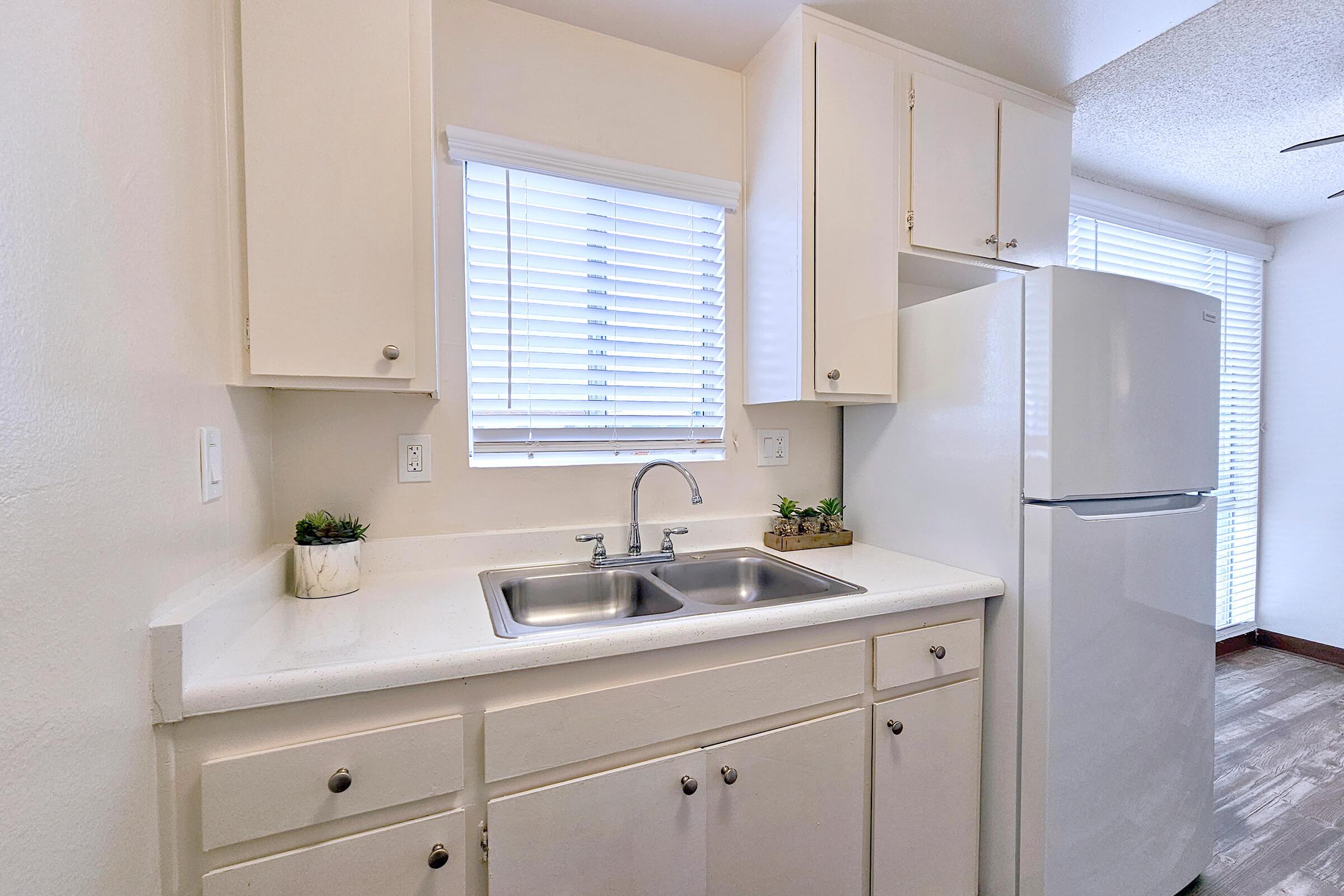 A clean, modern kitchen with a white countertop, double sink, and refrigerator. The cabinetry is light-colored and neatly arranged, with a window above the sink letting in natural light. Small potted plants are placed on the counter for a touch of greenery.