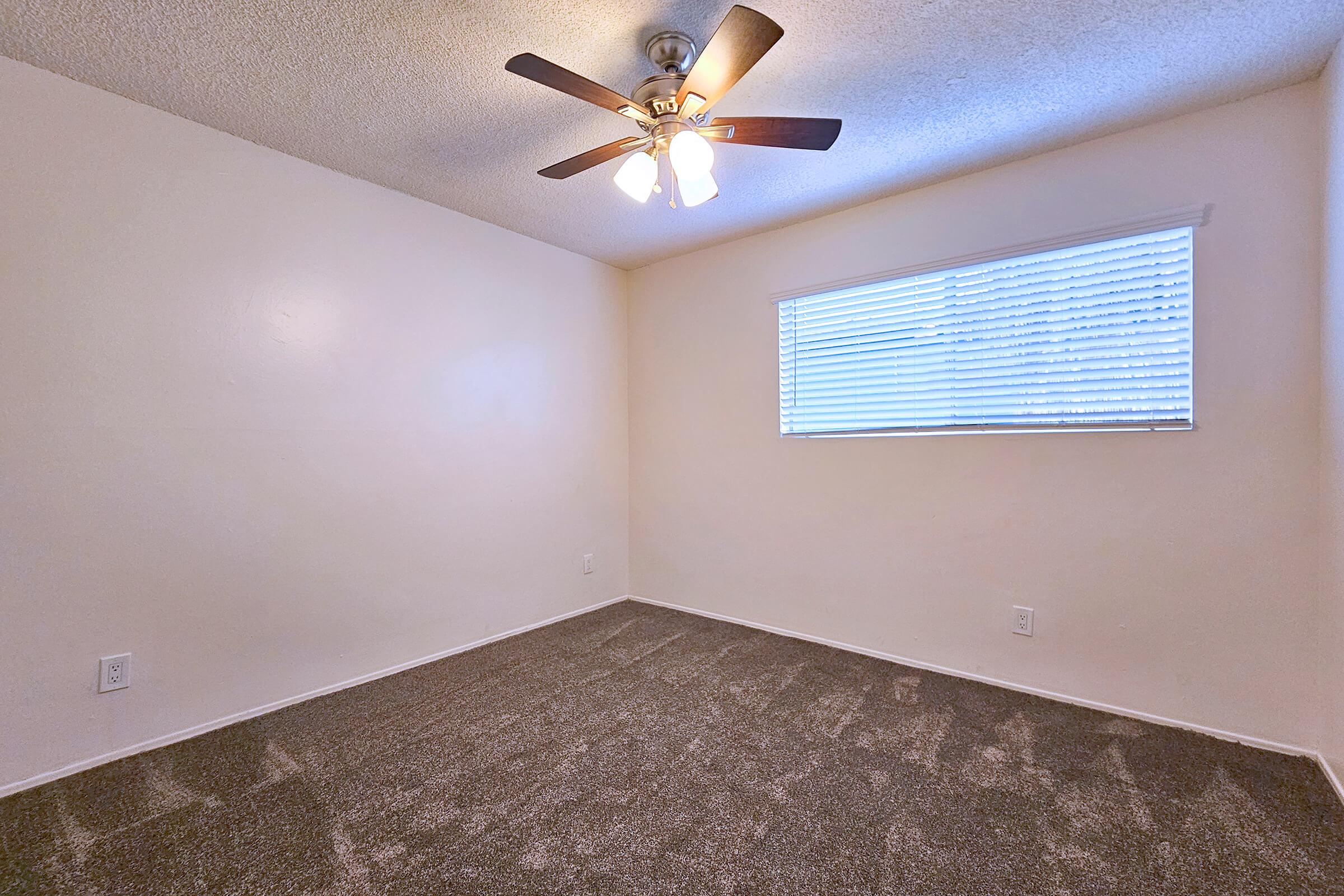 An empty room with light beige walls and a ceiling fan with four blades. There is a window on the right with white blinds, allowing natural light to enter. The floor is covered with gray carpet, and the overall atmosphere is bright and uncluttered.