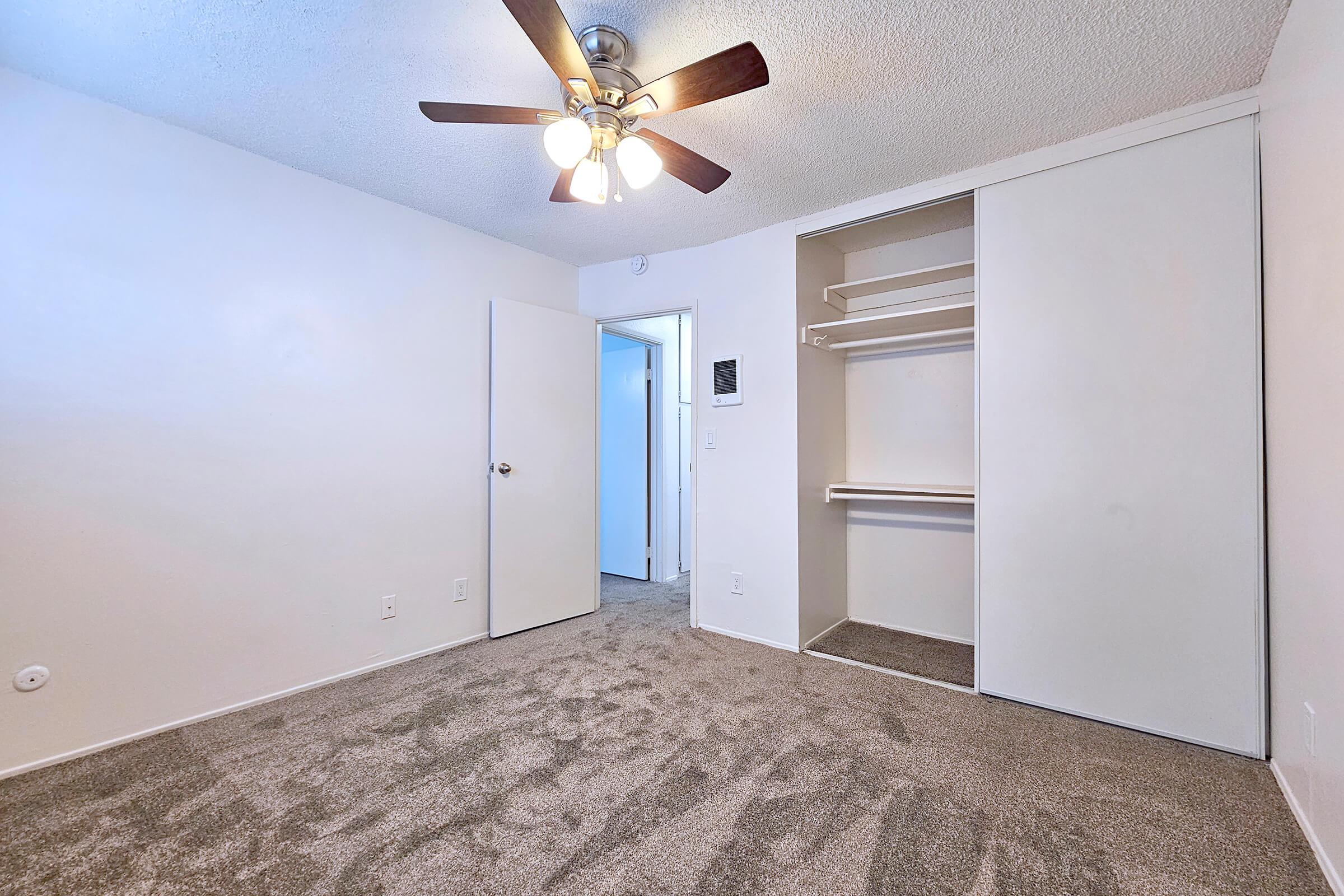 A well-lit room featuring a ceiling fan, beige carpet, and a closet with shelves. A closed door leads to another area, and the walls are painted white, creating a bright and spacious atmosphere.