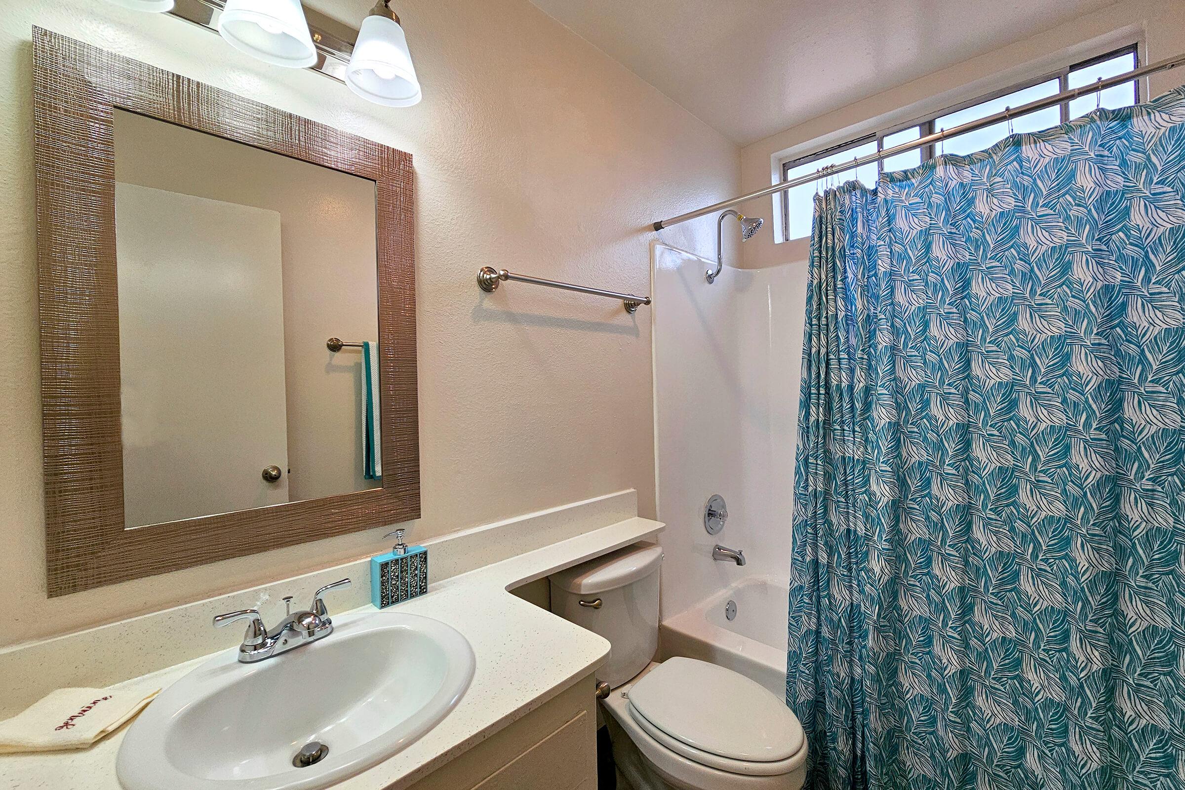 A bright and clean bathroom featuring a white sink with a modern faucet, a toilet, and a shower with a blue and white patterned curtain. A rectangular mirror is mounted above the sink, and natural light streams in through a window above the shower. Subtle decor complements the space.