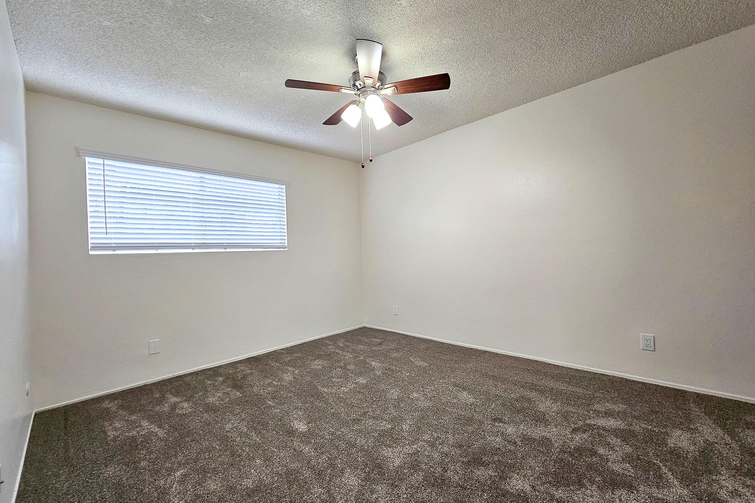 A well-lit, empty room with light gray carpet and white walls. There is a ceiling fan with wooden blades, and a window with white blinds allowing natural light to enter. The space is clean and uncluttered, suggesting it is ready for furnishing and personalization.