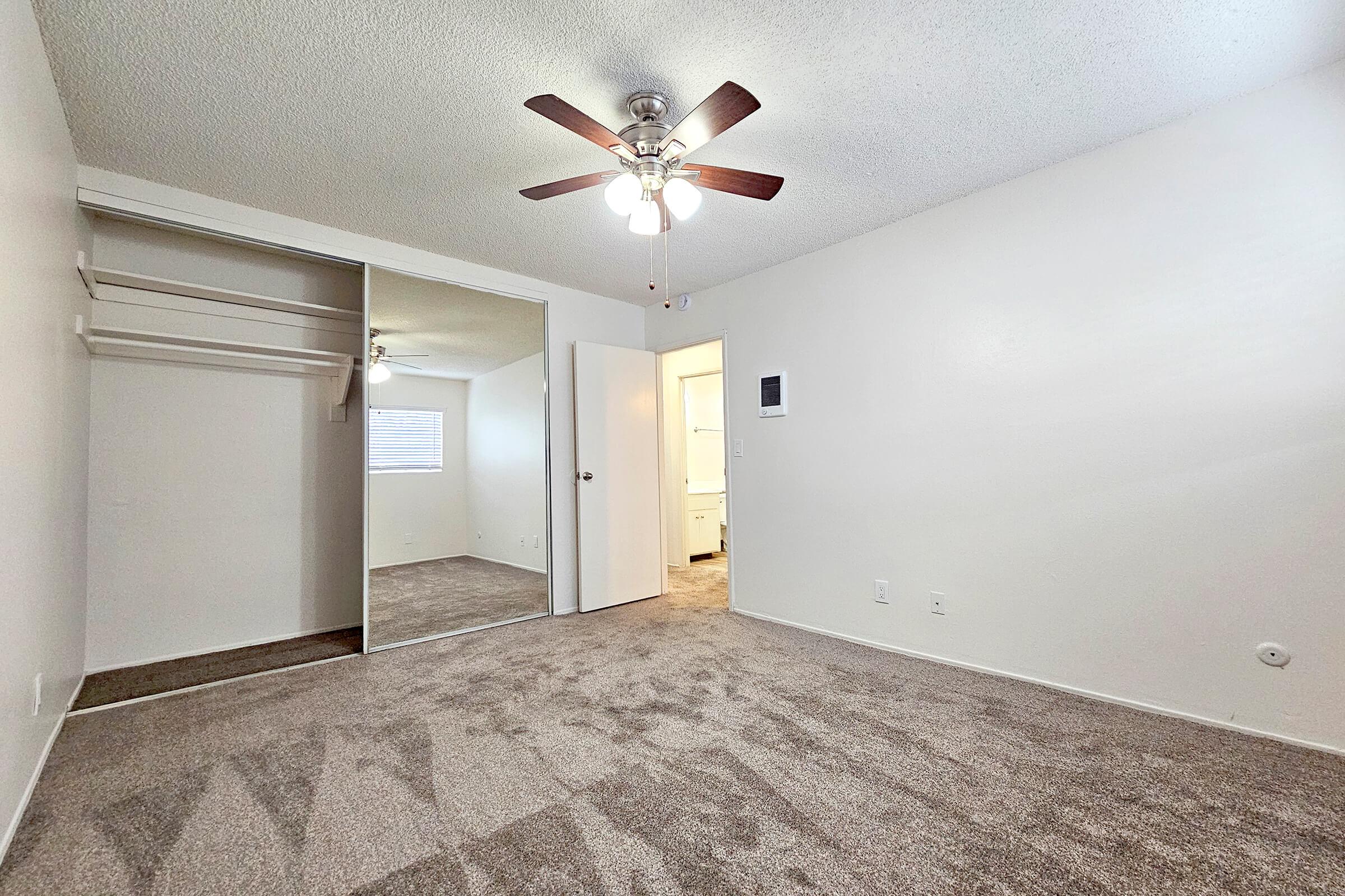 A spacious, empty room with light-colored walls and a ceiling fan. There's a closet on one side and a mirror reflecting the opposite wall. A door leads to another room, and natural light comes from a window. The carpeted floor is a soft shade, creating a warm and inviting atmosphere.