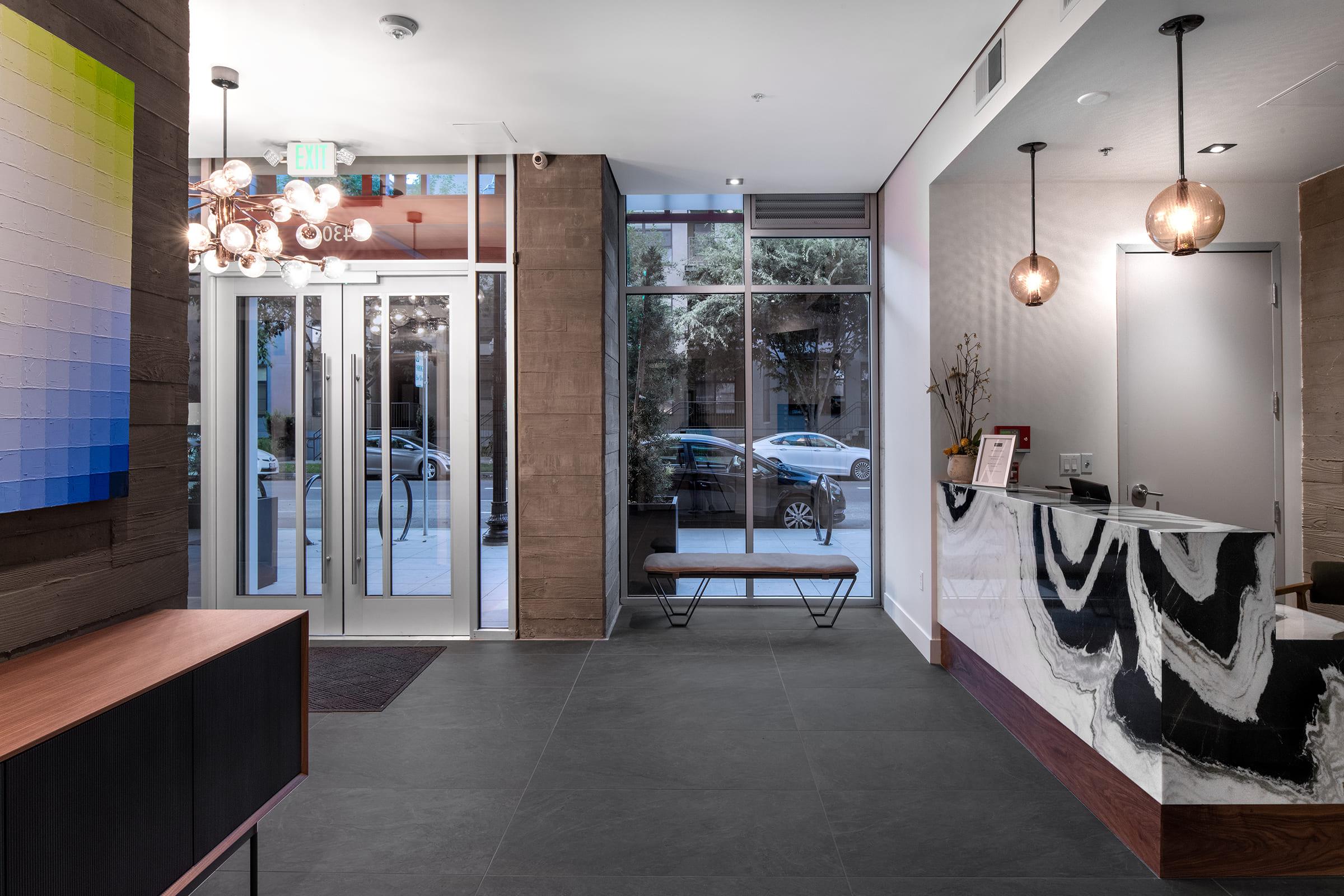 Modern lobby interior featuring a sleek reception desk with a marble front, stylish light fixtures, large glass doors leading outside, and a minimalist seating area. A colorful abstract artwork is displayed on the wall, and natural light illuminates the space, creating an inviting atmosphere.