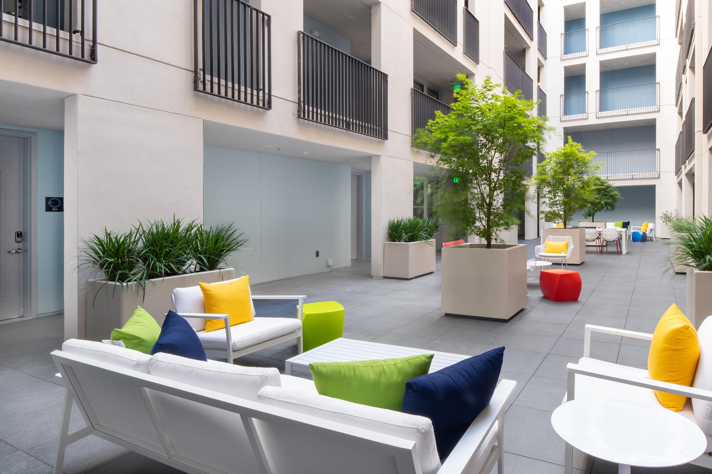 A modern courtyard featuring comfortable seating areas with white sofas adorned with colorful cushions in yellow, green, blue, and red. Potted plants and small trees line the space, surrounded by a contemporary building with balconies. Natural light enhances the inviting atmosphere.