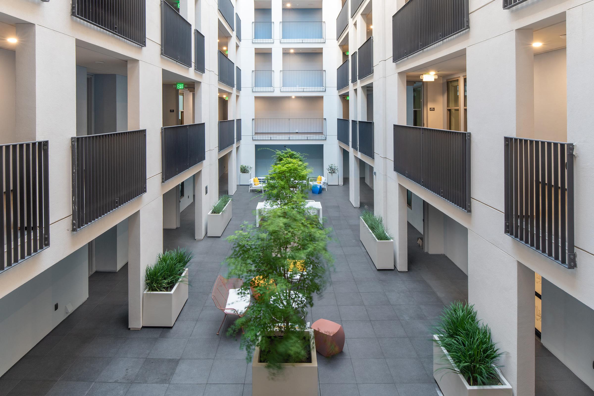 A modern, open courtyard featuring greenery and contemporary furniture. The space is surrounded by multi-story buildings with balconies, creating a bright and inviting atmosphere. Potted plants and seating areas provide a relaxing environment in an urban setting.