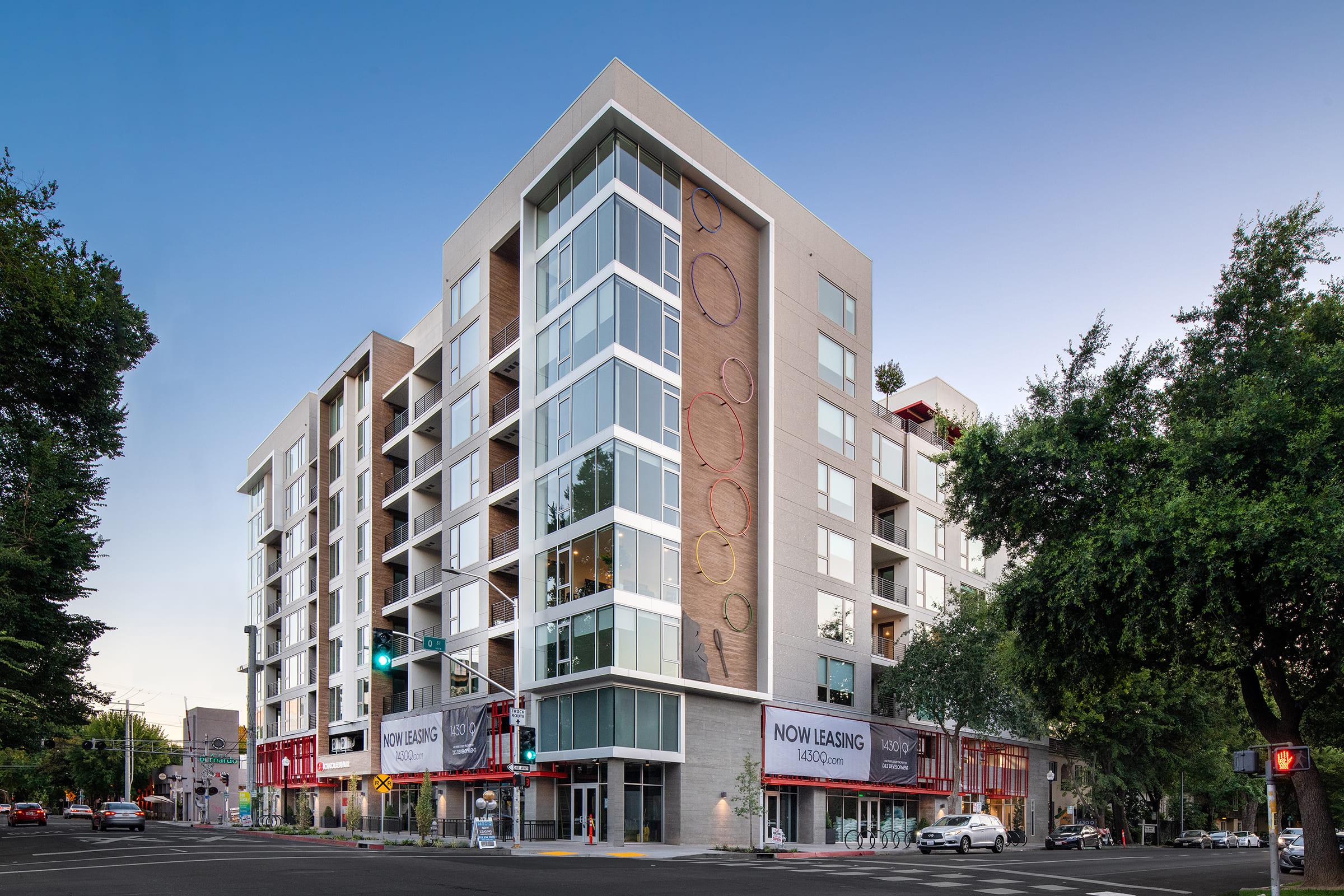 A modern multi-story apartment building with large windows, featuring a sleek design. The ground floor includes retail space with "NOW LEASING" signs visible. Surrounding the building are trees and urban elements, set against a blue sky.