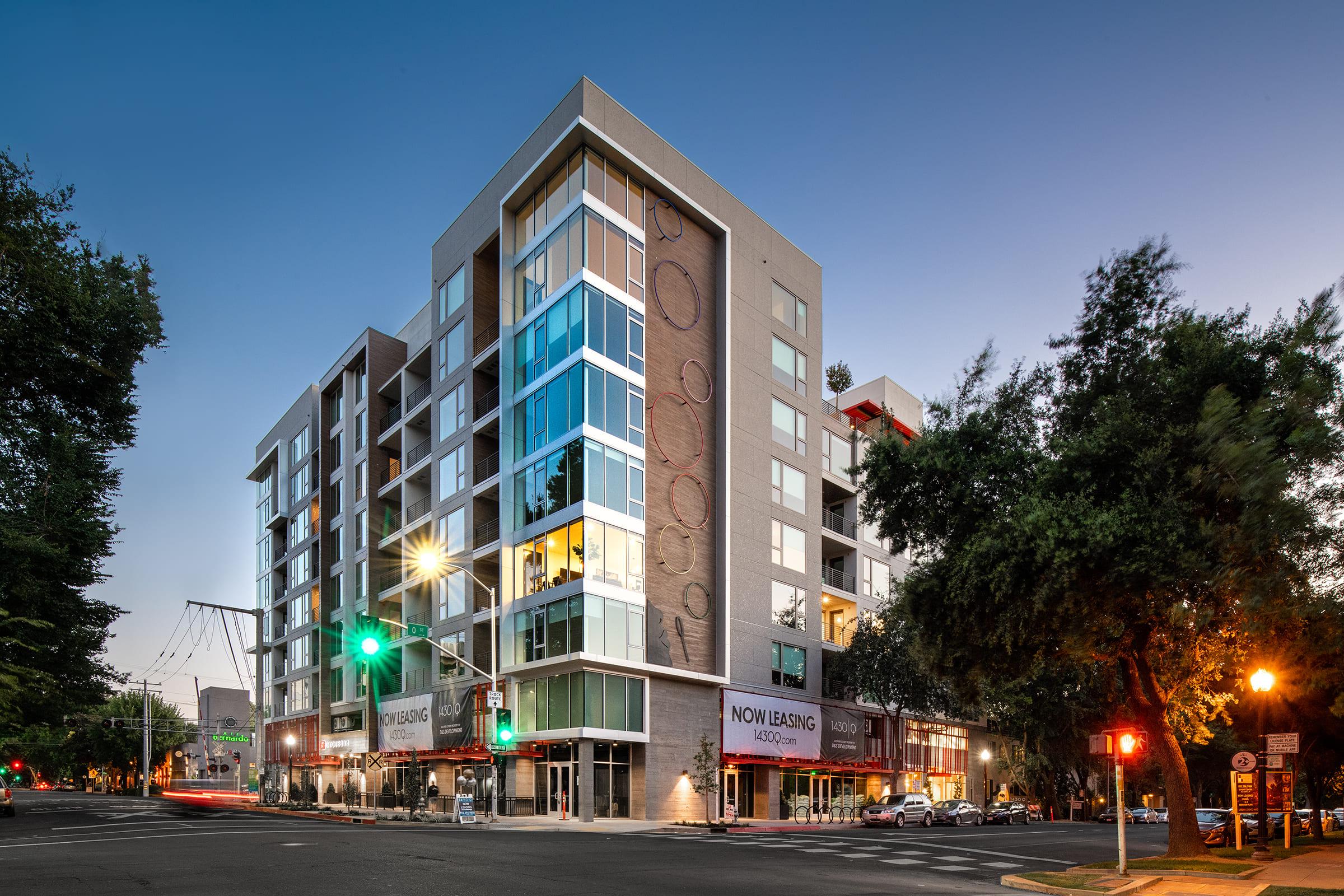 Modern multi-story building with large glass windows and a contemporary design, located at a street intersection. The structure features colorful accents and banners advertising available rentals. Trees and streetlights are visible in the surrounding area during twilight.