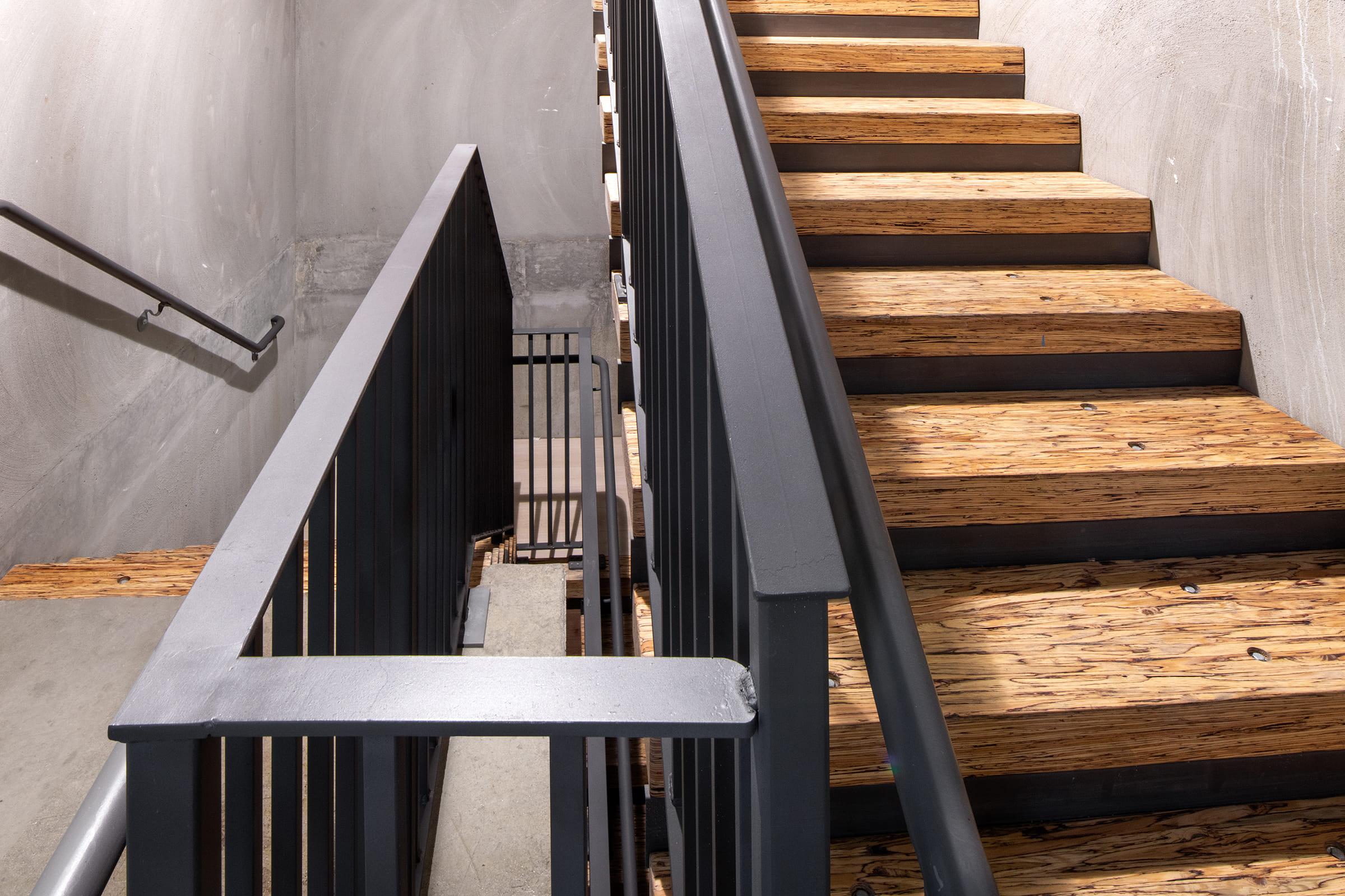 Staircase with wooden steps and a metal railing, leading upwards. The wall is bare concrete, and the design is modern and minimalistic. The scene highlights the texture of the wooden steps and the clean lines of the railing.