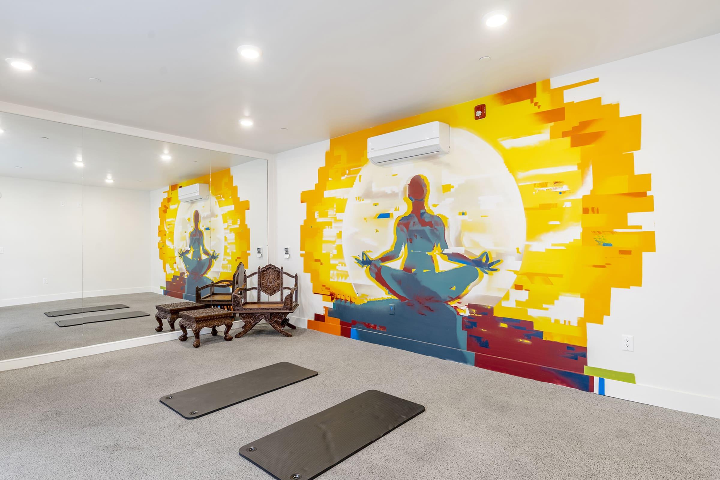 A bright, modern yoga studio featuring a large mural of a meditating figure against a vibrant, sunlit background. The room includes two yoga mats on the floor and a decorative wooden chair. Mirrors on one wall enhance the sense of space and light