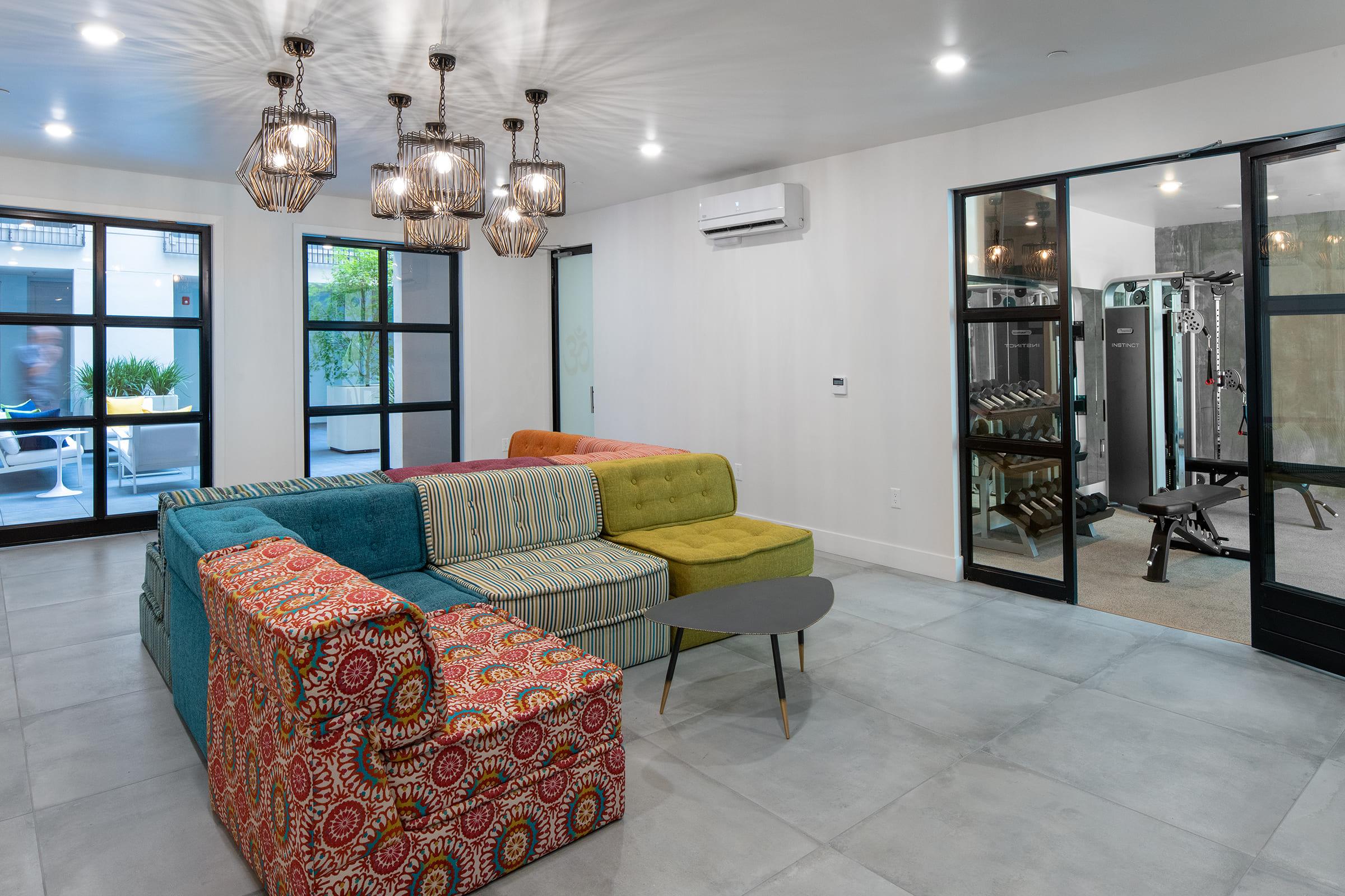 A modern lounge area featuring a multi-colored sectional sofa with various patterns, a small round table, and stylish pendant lighting. Large windows provide natural light, revealing a glimpse of an adjacent gym area through glass doors. The flooring is light gray tile, creating a contemporary ambiance.