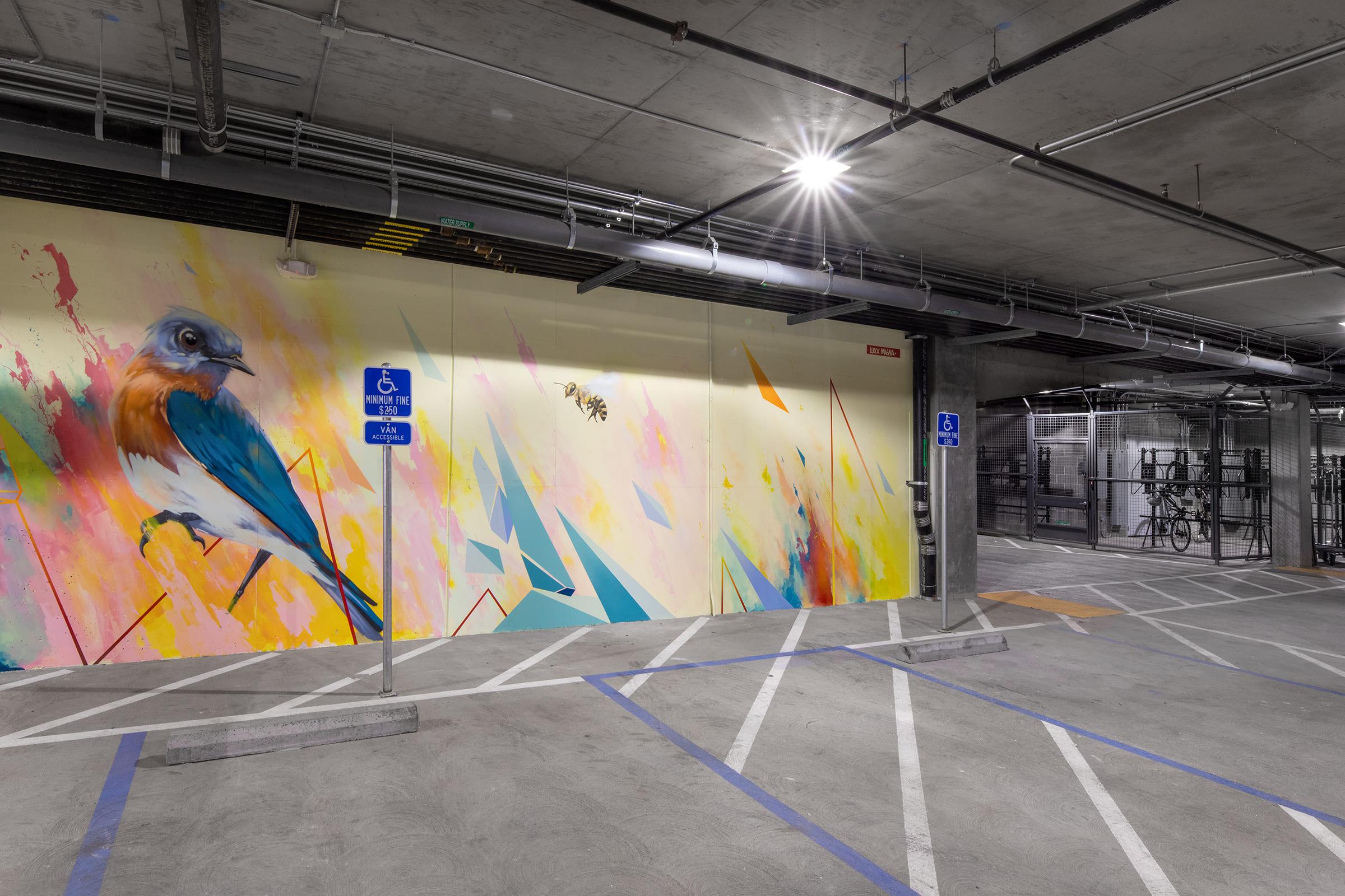 A modern parking garage featuring a vibrant mural of a colorful bird against an abstract background of geometric shapes. The area includes designated parking spaces, including those for handicapped access, with bike racks visible in the background. The lighting is bright and industrial.