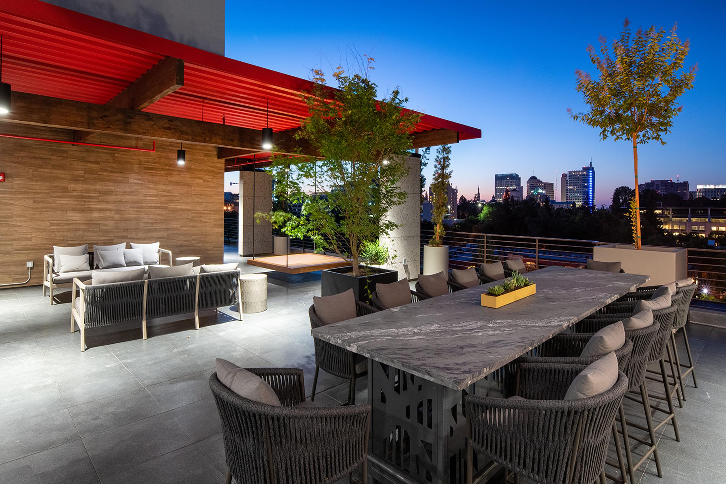 A modern rooftop patio showcasing a large dining table surrounded by stylish chairs. Comfortable seating areas are arranged with plush cushions, and greenery adds a touch of nature. The scene is set against a twilight sky, with city buildings illuminated in the background.