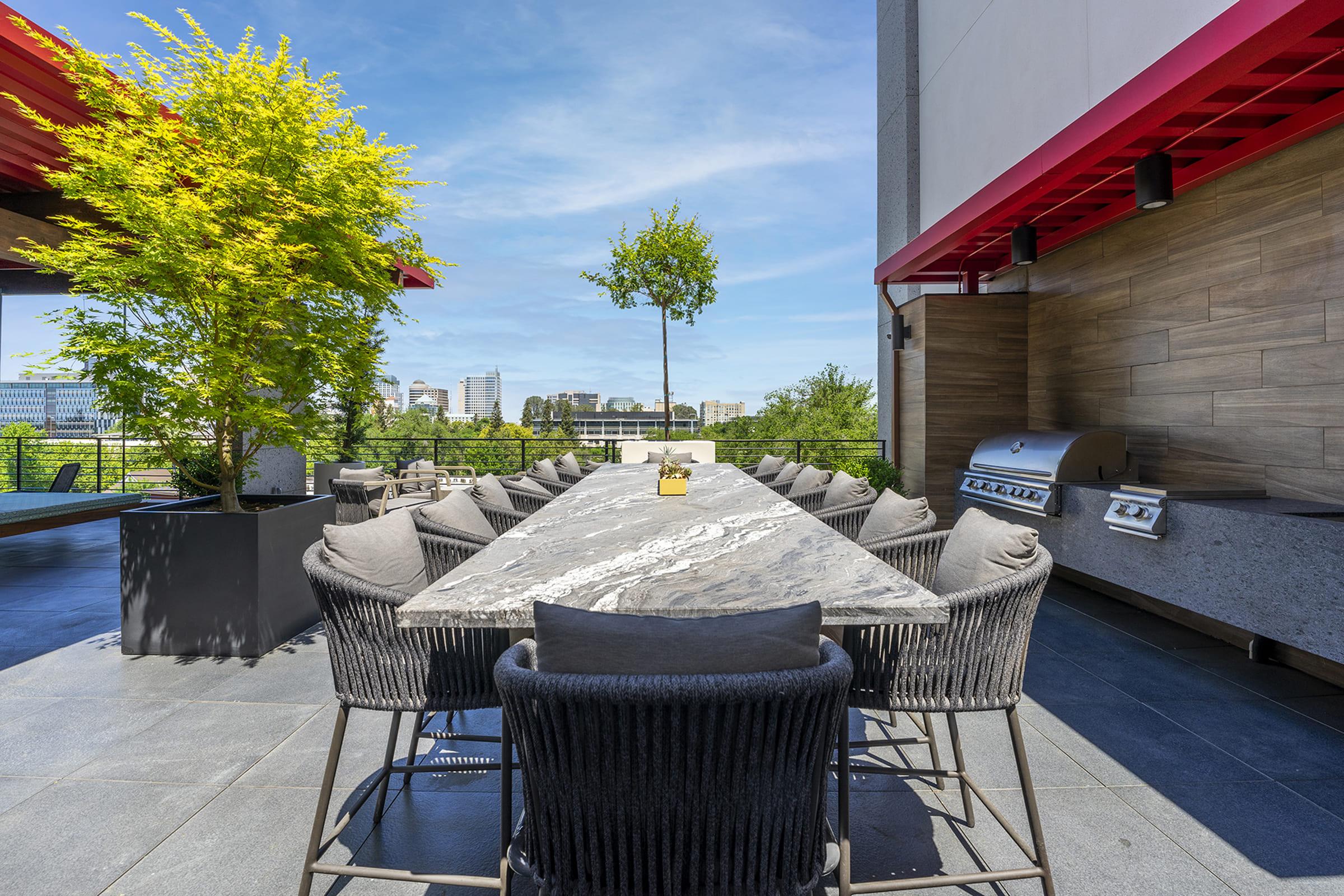A spacious outdoor dining area featuring a long marble table surrounded by stylish black chairs. Lush greenery and a small tree enhance the setting, with a modern grill visible in the background against a clear blue sky. Ideal for gatherings and enjoying fresh air.