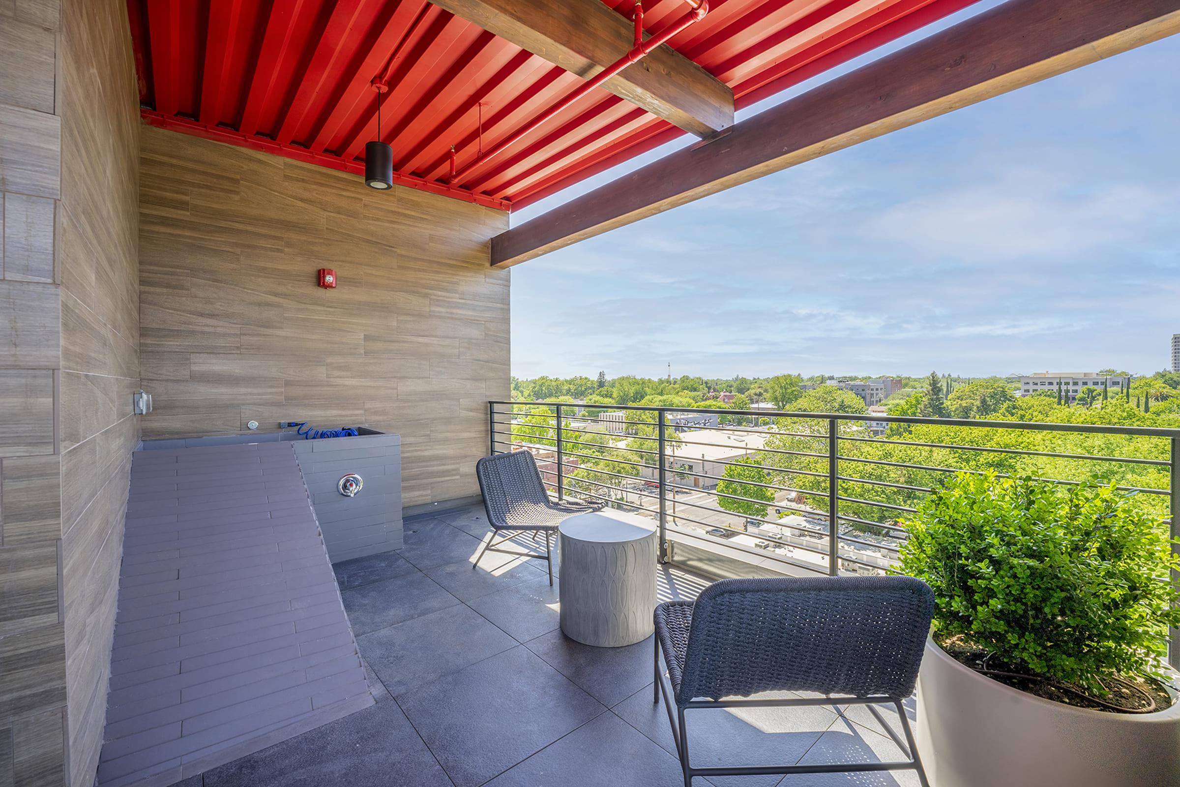 A modern balcony with two chairs and a small table, surrounded by greenery. The floor is tiled, and there's a railing with a view of trees and buildings in the background. A storage unit is partially visible on the left, and the ceiling has exposed beams and red steel. Bright day with clear skies.