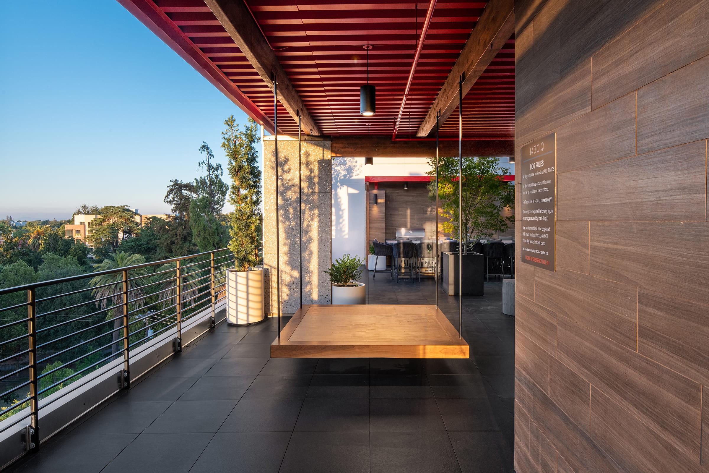 A spacious outdoor balcony featuring a wooden swing, sleek tile flooring, and railing. Lush greenery and distant hills are visible in the background under a clear blue sky. Modern furniture and a dining area can be seen, emphasizing a stylish and inviting atmosphere.