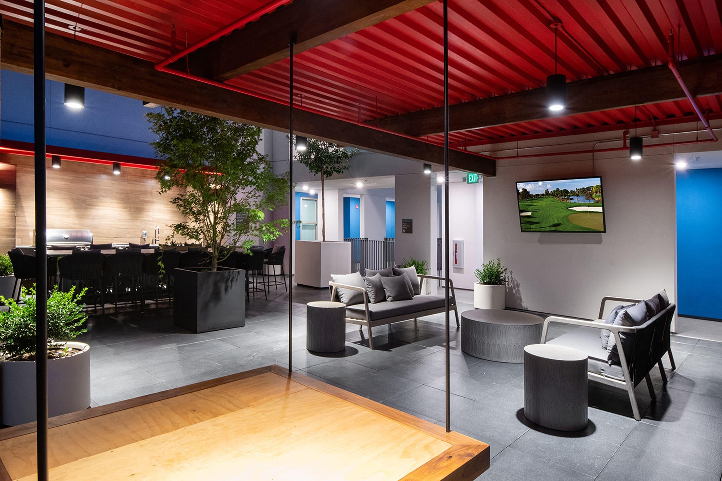 Modern indoor lounge area featuring a mix of seating options, including couches and round tables. A large television displays a golf course scene. The space is bright with a red ceiling, wood accents, and greenery, creating a relaxed atmosphere for gatherings and social interactions.