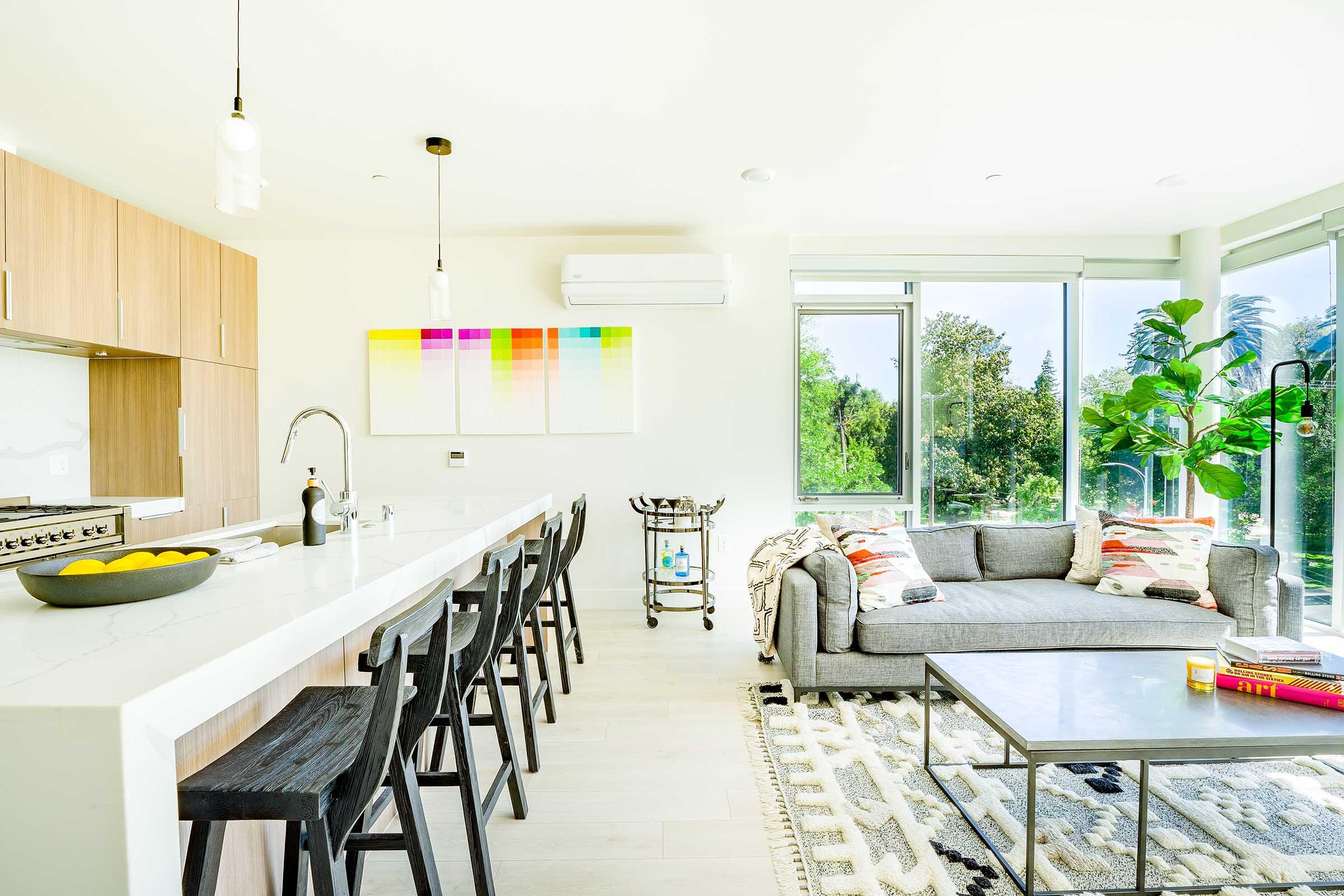 A bright, modern kitchen and living area featuring a sleek countertop with bar stools, a large window with a view of greenery, a cozy gray sofa adorned with colorful pillows, a coffee table, and decorative plants. A colorful wall art piece enhances the airy atmosphere.