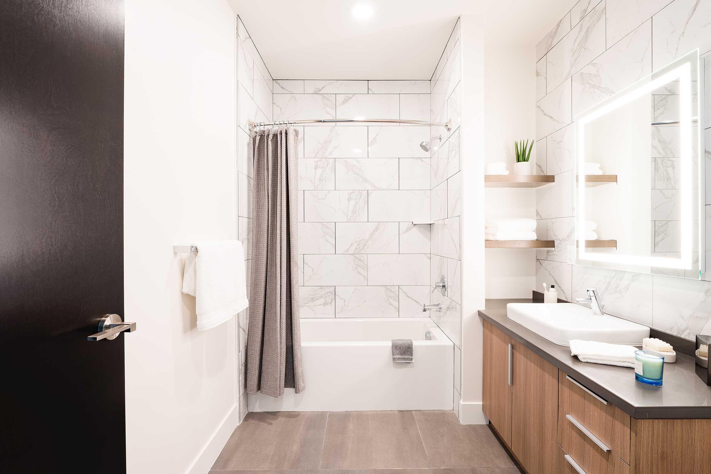 Modern bathroom featuring a bathtub with a shower, gray and white tiled walls, a sleek wooden vanity with a sink, and shelves with neatly folded towels. A large mirror with built-in lighting is mounted above the sink, and a door leads to the hallway. Soft, neutral colors create a calming ambiance.