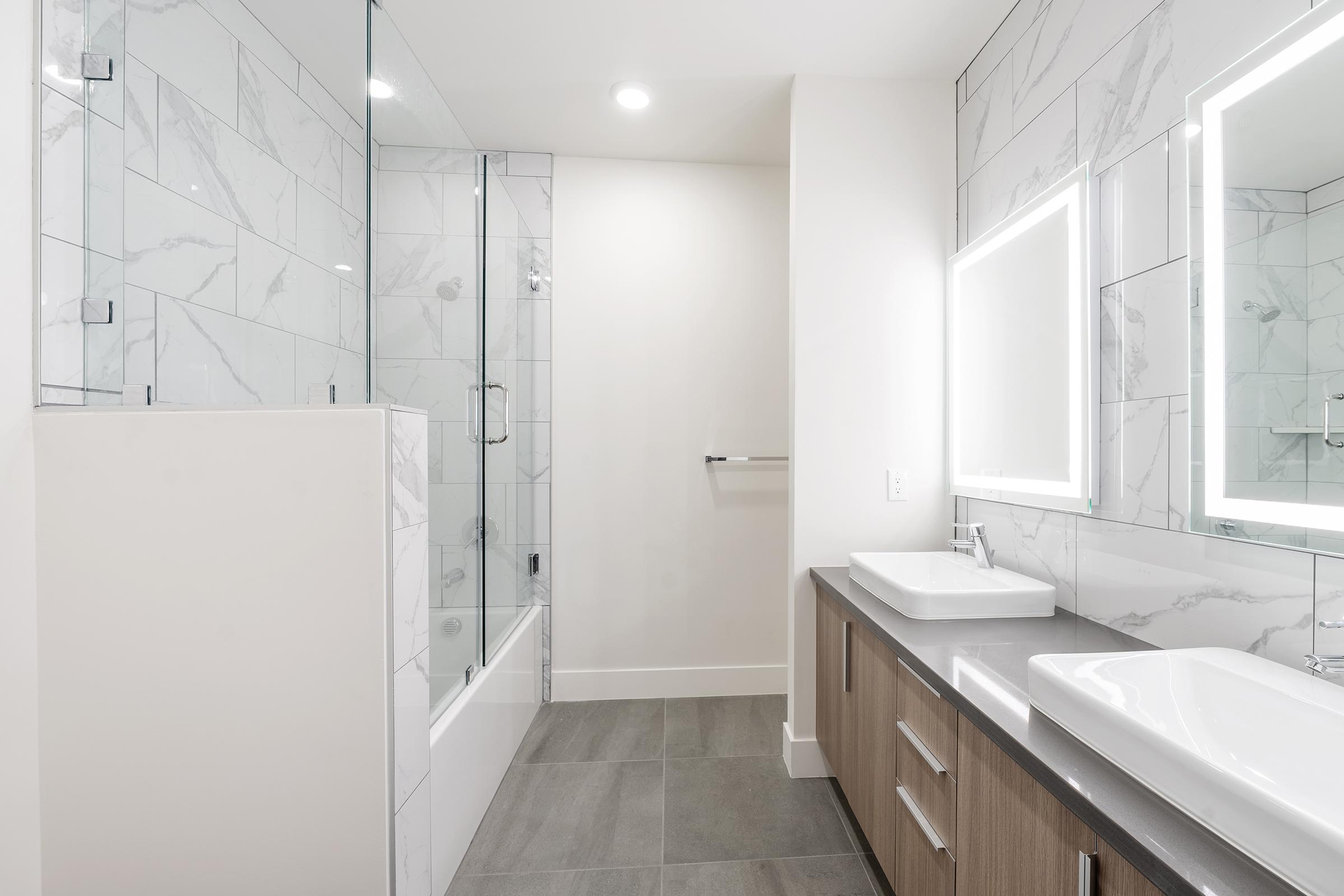 A modern bathroom featuring a glass shower enclosure, a bathtub, two rectangular sinks with sleek faucets, and backlit mirrors. The walls are adorned with white marble tiles, and the floor is a neutral-colored tile. Bright lighting enhances the clean and spacious design.
