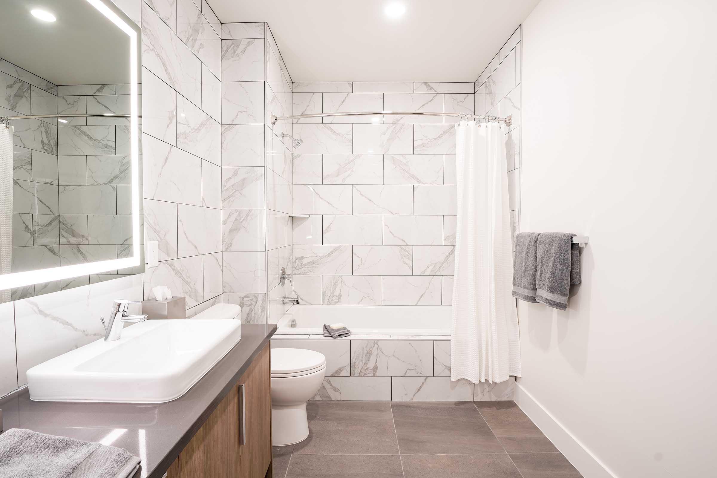 A modern bathroom featuring white marble tile walls, a bathtub with a shower curtain, a sleek sink with a mirror, and wooden cabinetry. Gray towels are displayed on a rack, and soft lighting enhances the clean, contemporary design.