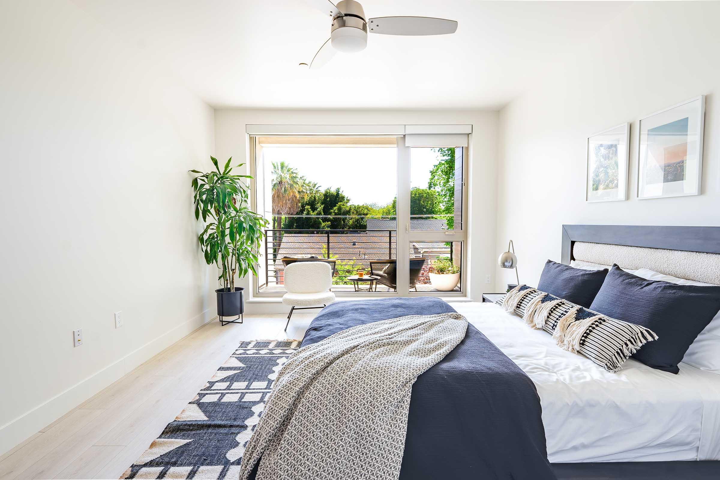 A modern bedroom featuring a large bed with dark bedding and decorative pillows. There’s a stylish chair next to a sliding glass door that leads to a balcony with greenery visible outside. A potted plant and a floor lamp add a touch of design, while a patterned rug lies on the floor, enhancing the cozy atmosphere.