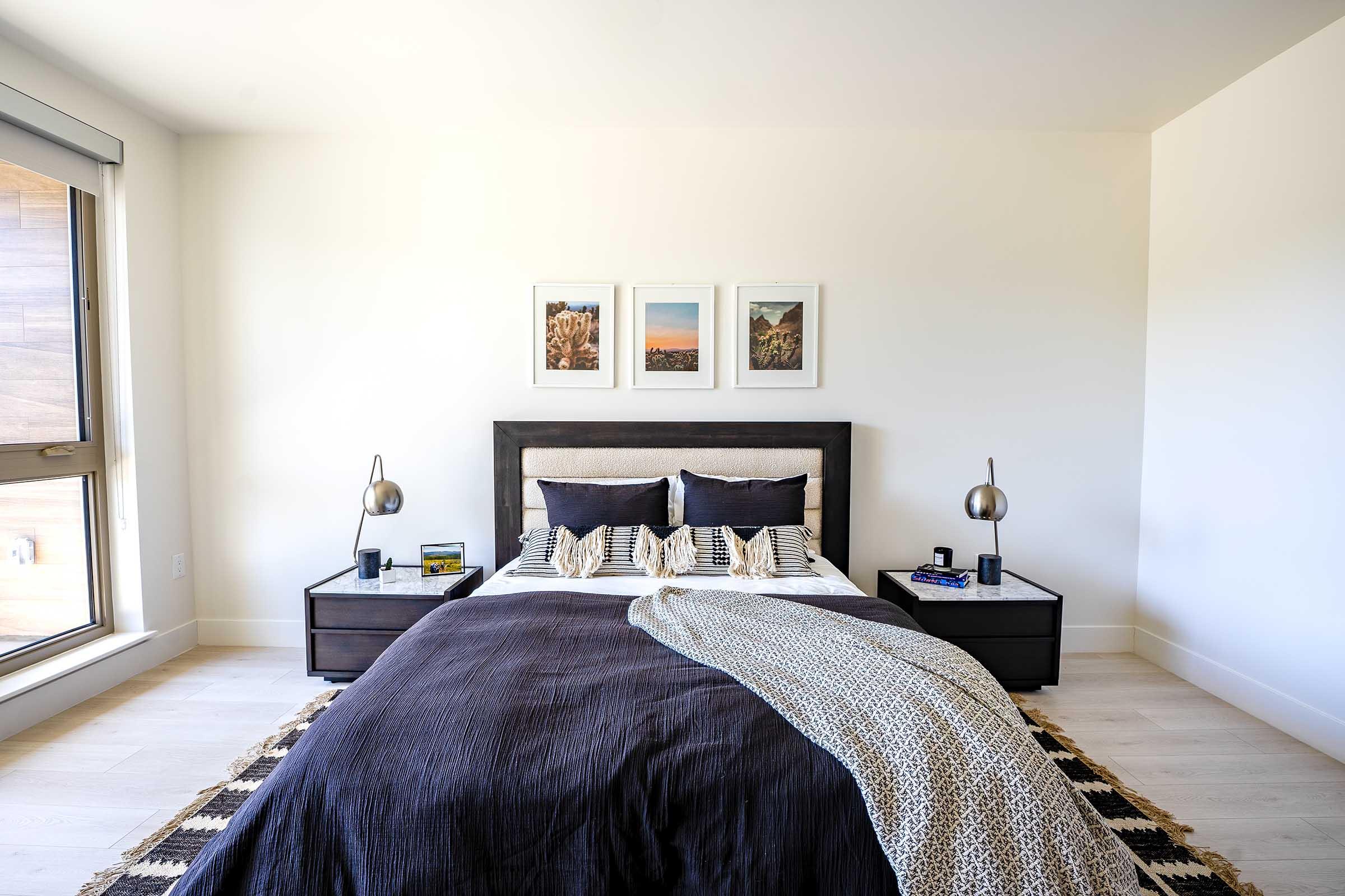 A modern bedroom featuring a large bed with dark bedding and patterned throw pillows. Two bedside tables with lamps flanking the bed, and three framed pictures hang above the headboard. A large window lets in natural light, and a patterned rug is placed on the light-colored wooden floor.
