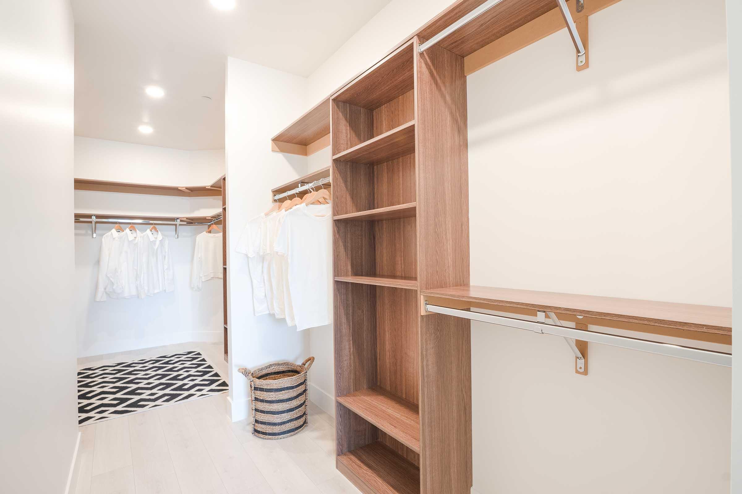 A spacious, organized closet featuring wooden shelves, hanging racks with white clothing, a woven basket, and a black and white patterned rug on the floor. The walls are light-colored, and the space is brightly lit with ceiling lights.