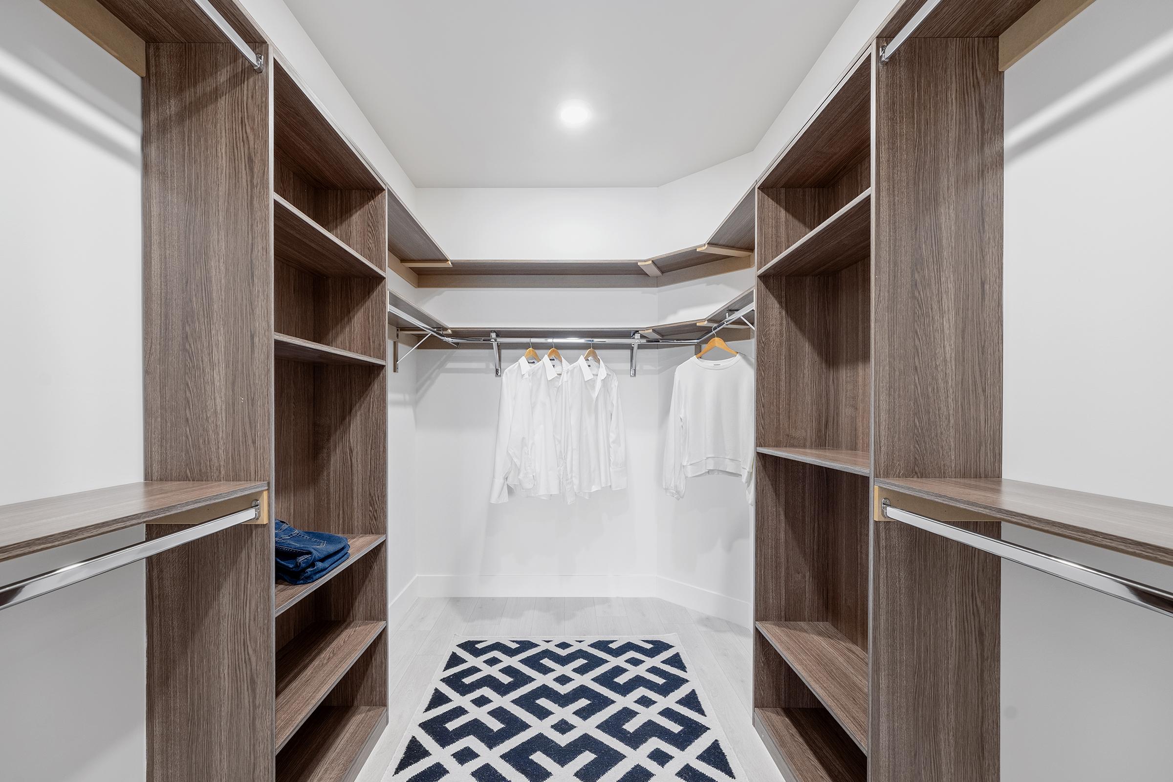 A spacious walk-in closet featuring wooden shelves and hanging rods. Two white shirts are neatly hung on hangers, while folded blue jeans rest on the shelf. The floor is covered with a geometric patterned rug, and the walls are painted white, creating a bright and organized space.