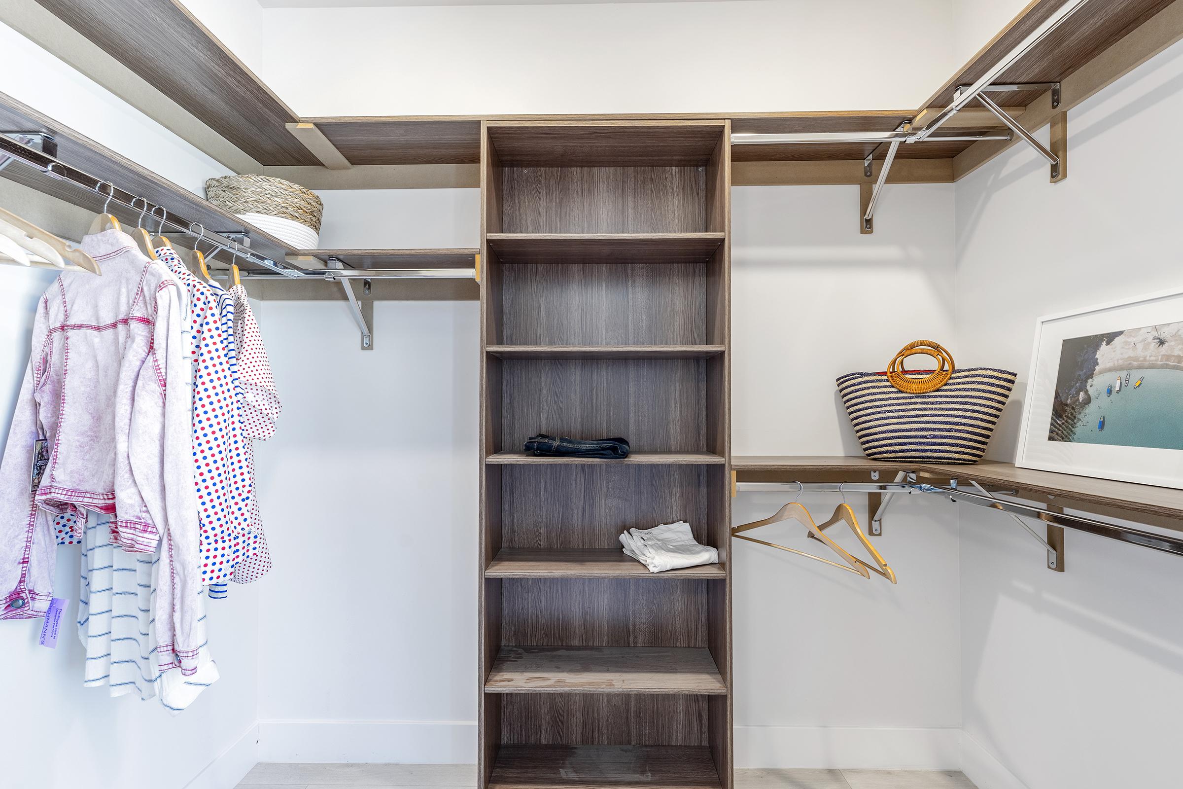 A well-organized walk-in closet featuring wooden shelves and hanging rods. On the left side, there are neatly hung clothes in various patterns, while the right side has decorative items, including a striped basket and a framed picture. Empty shelf space is available for additional storage.