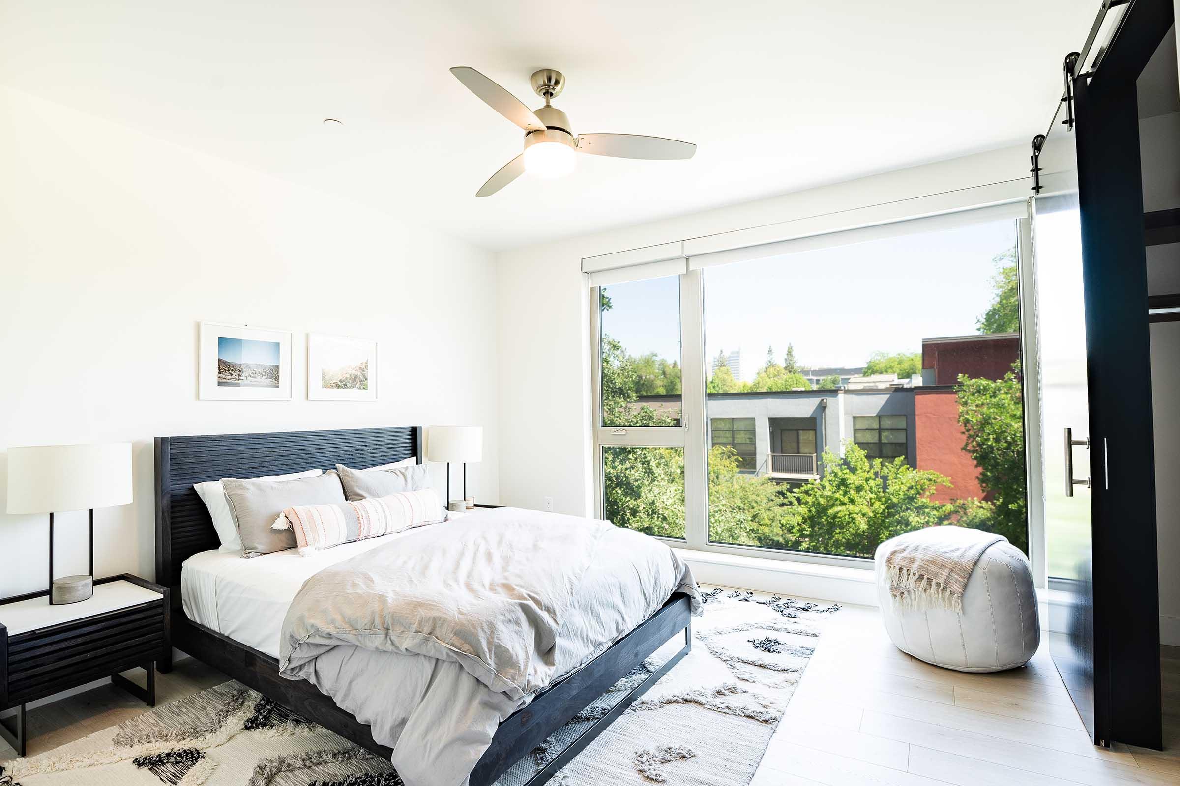 A bright and airy bedroom featuring a large window with outdoor views, a comfortable bed with light gray bedding, and modern furnishings including nightstands with lamps. A cozy pouf sits on the floor, and framed pictures adorn the walls, enhancing the contemporary aesthetic of the space.